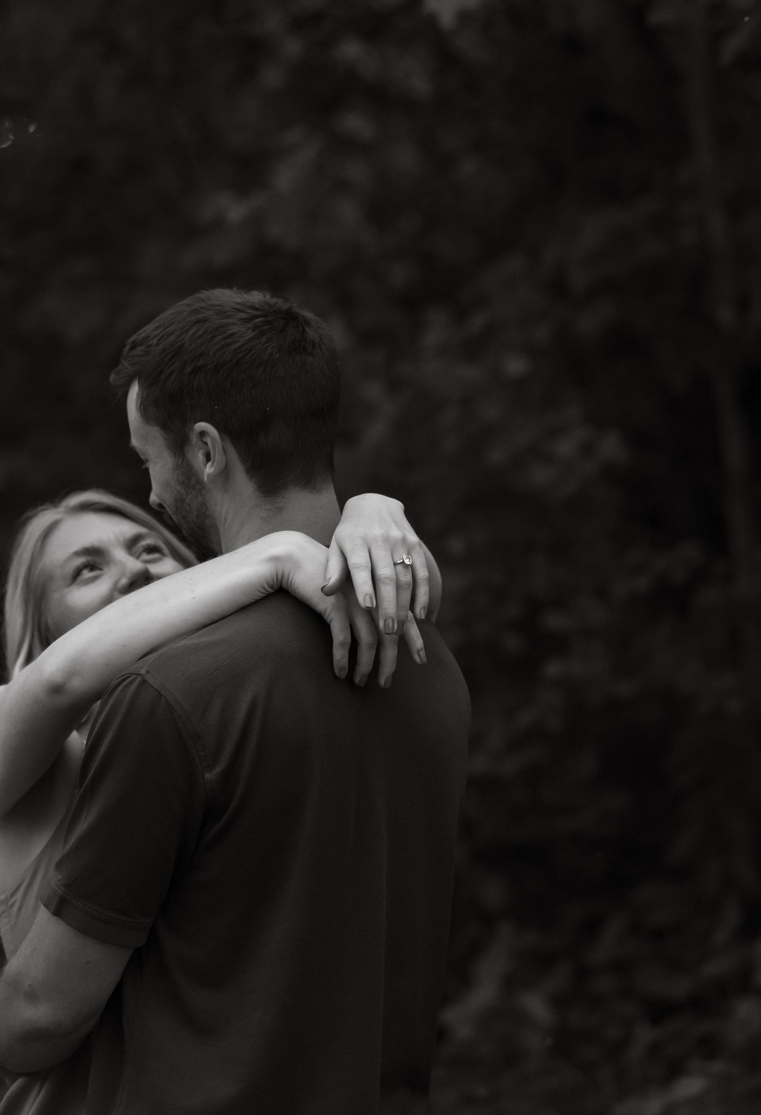 Black and white photo of a couple embracing outdoors, woman resting her head on man's shoulder showing engagement ring.