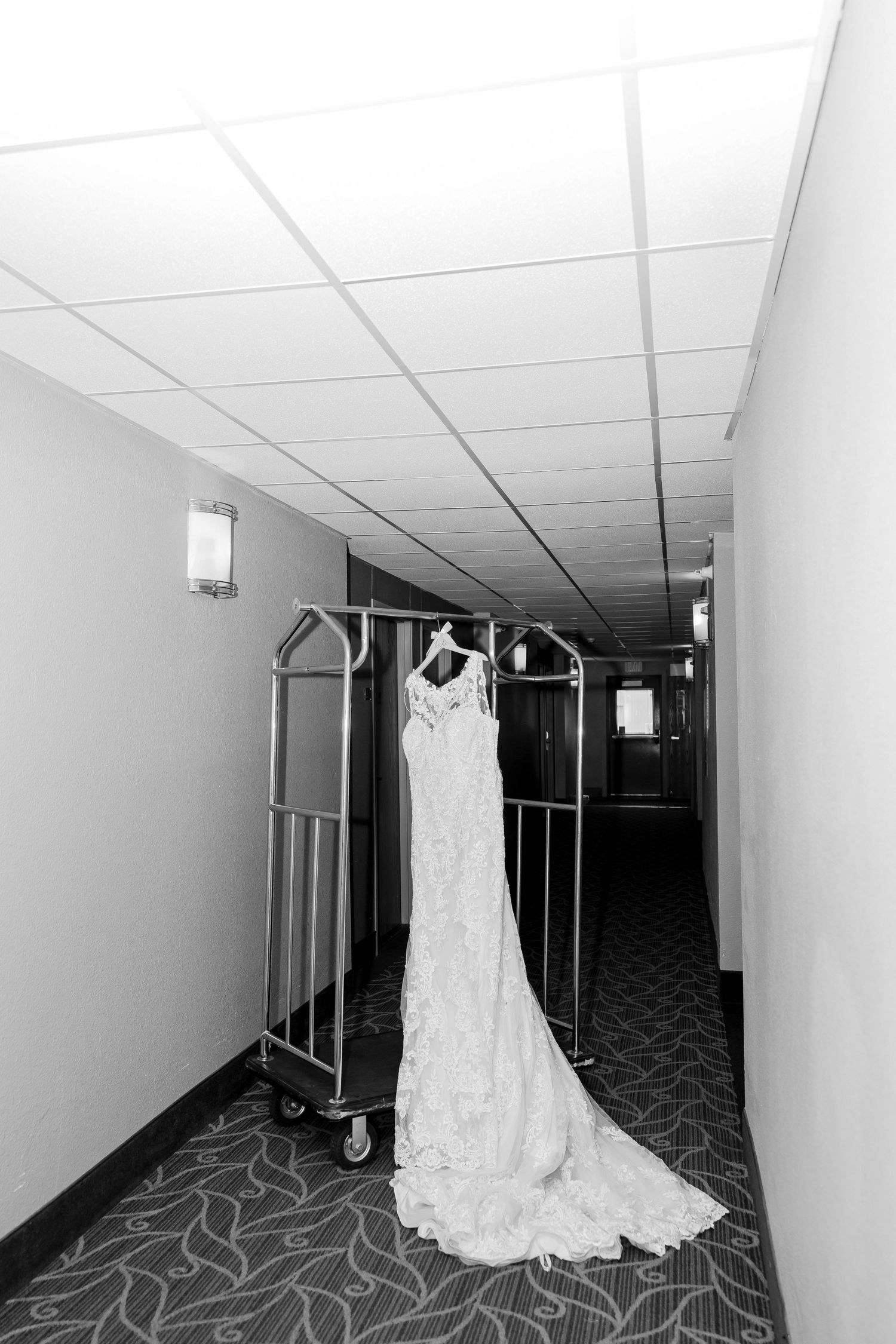 Wedding dress hangs in empty hotel hallway creating dramatic black and white composition.