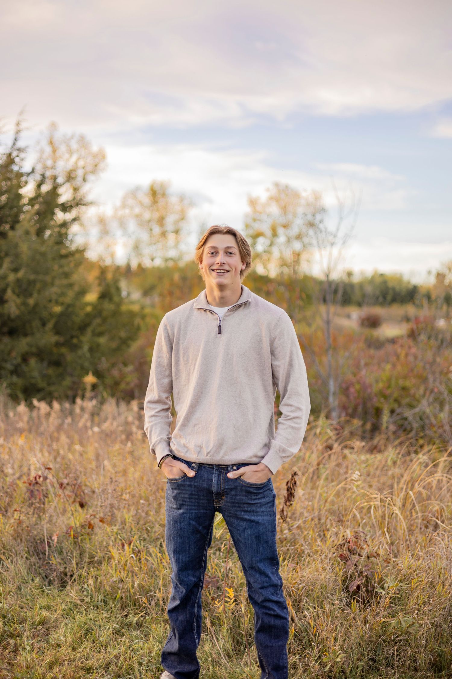 Person in beige hoodie and jeans poses casually in field during golden hour.