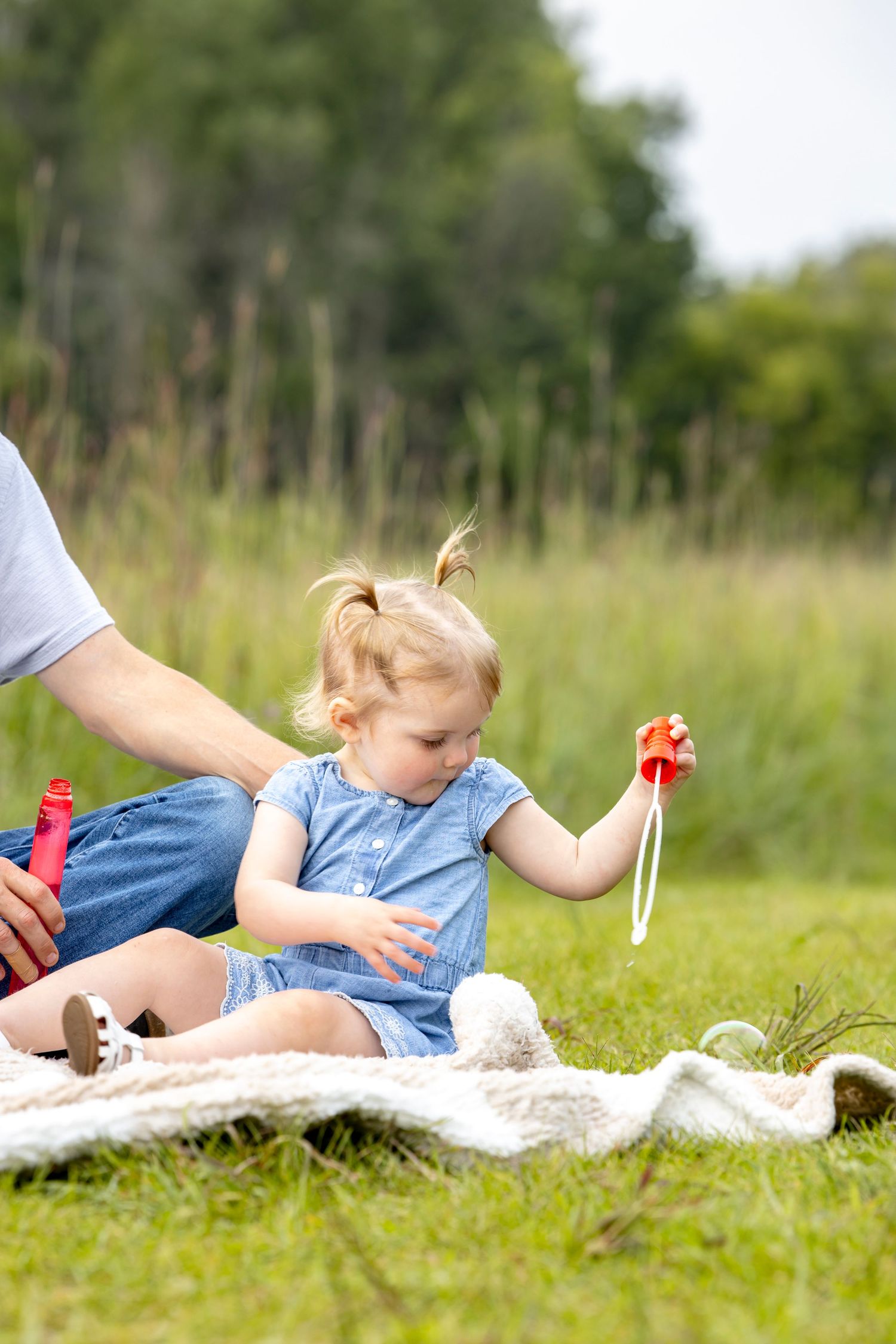 Child in blue dress playing with bubbles while lying on white blanket outdoors.