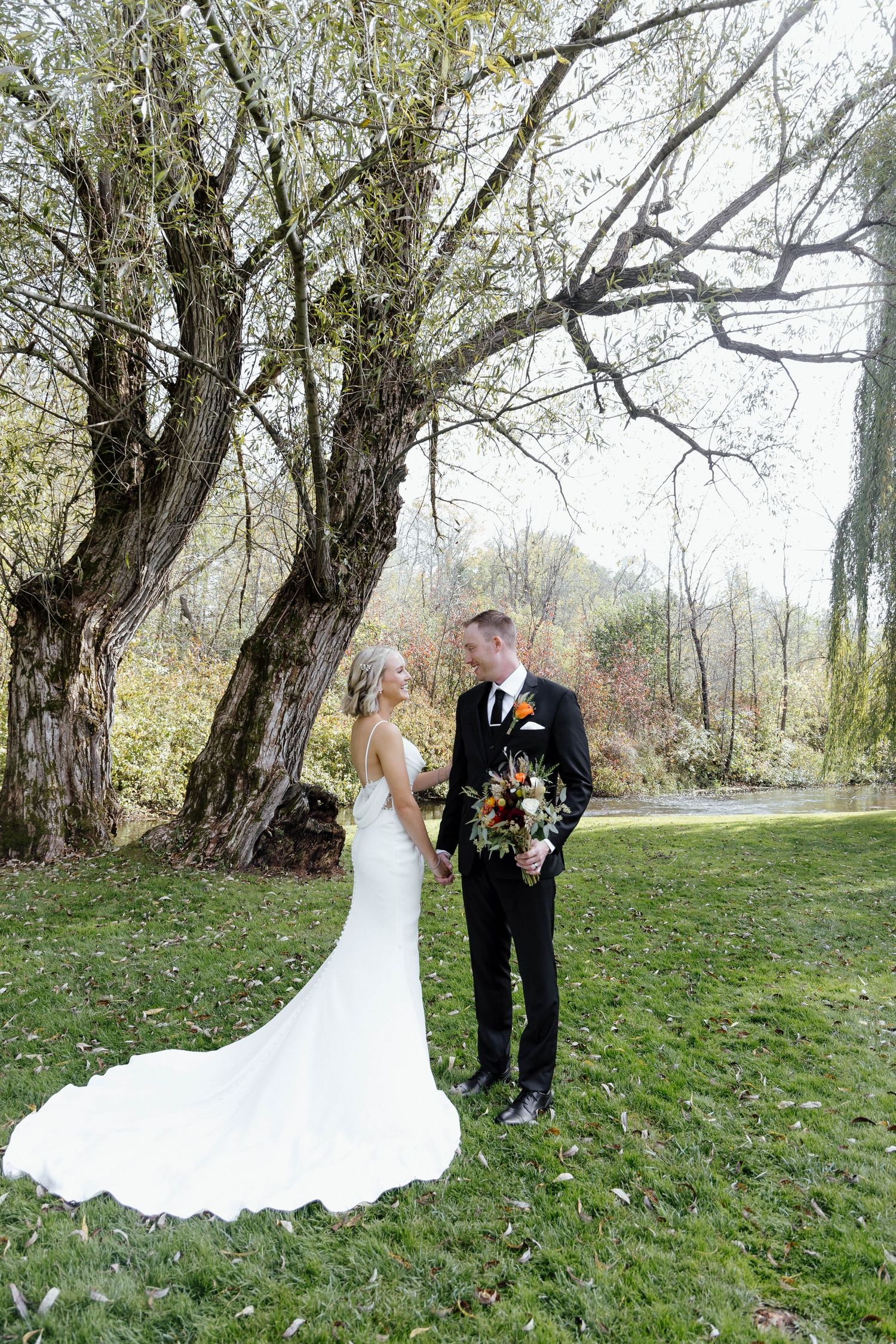 Wedding couple poses among autumn trees on grassy hillside with flowing white dress.