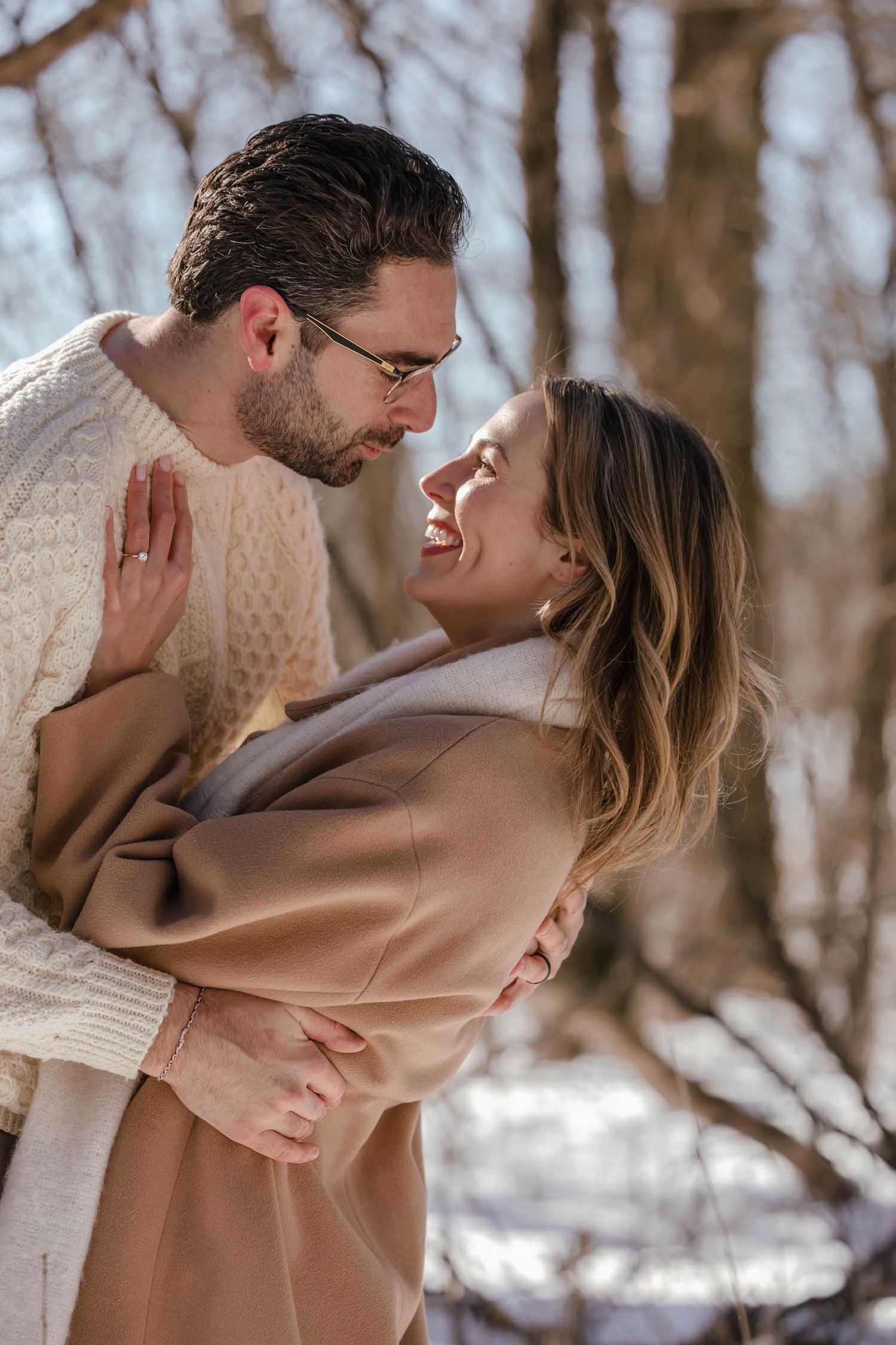 A romantic winter moment between a couple in cream and camel colored outfits among snowy trees.