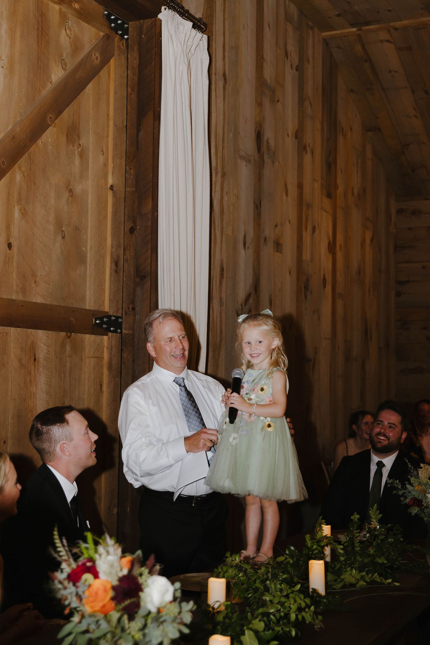 A celebration speech being given at a rustic barn wedding reception with floral table decorations.