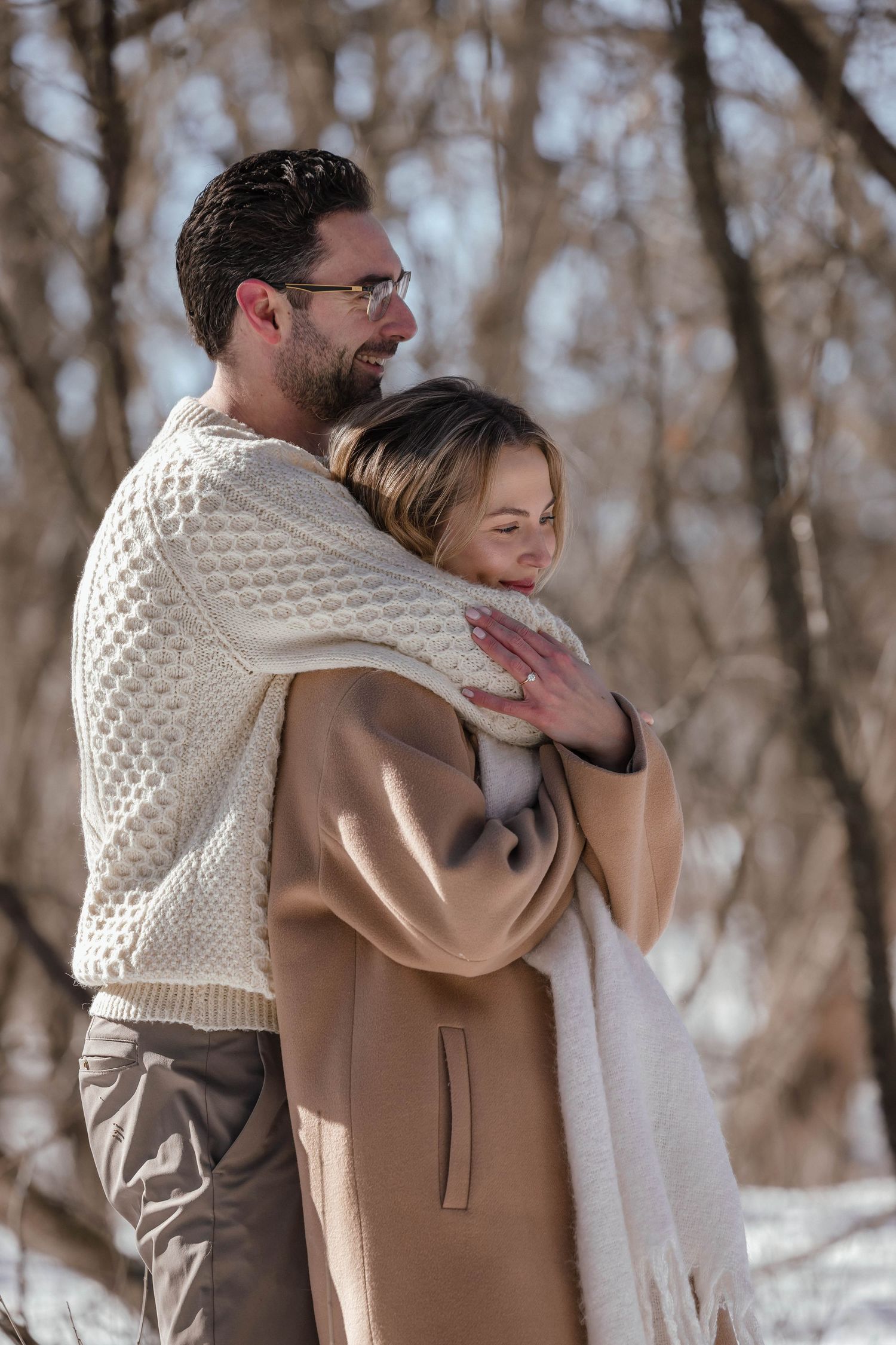 A warm embrace in the snow with one person in a cream sweater and another in a camel coat.