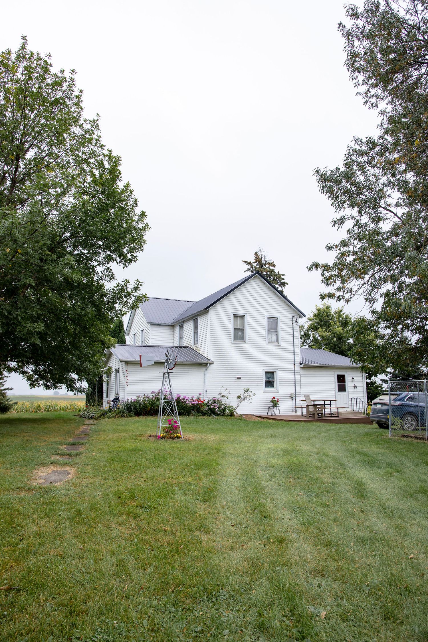White farmhouse with windmill and lawn set against a cloudy sky in a rural setting.