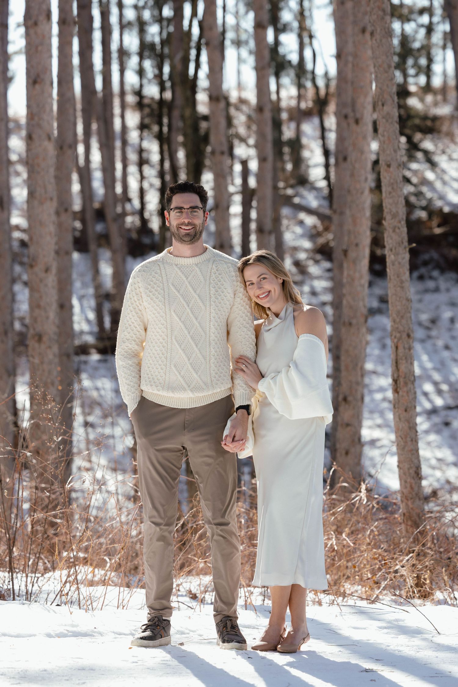 A couple poses together in a snowy forest wearing white and beige winter outfits.