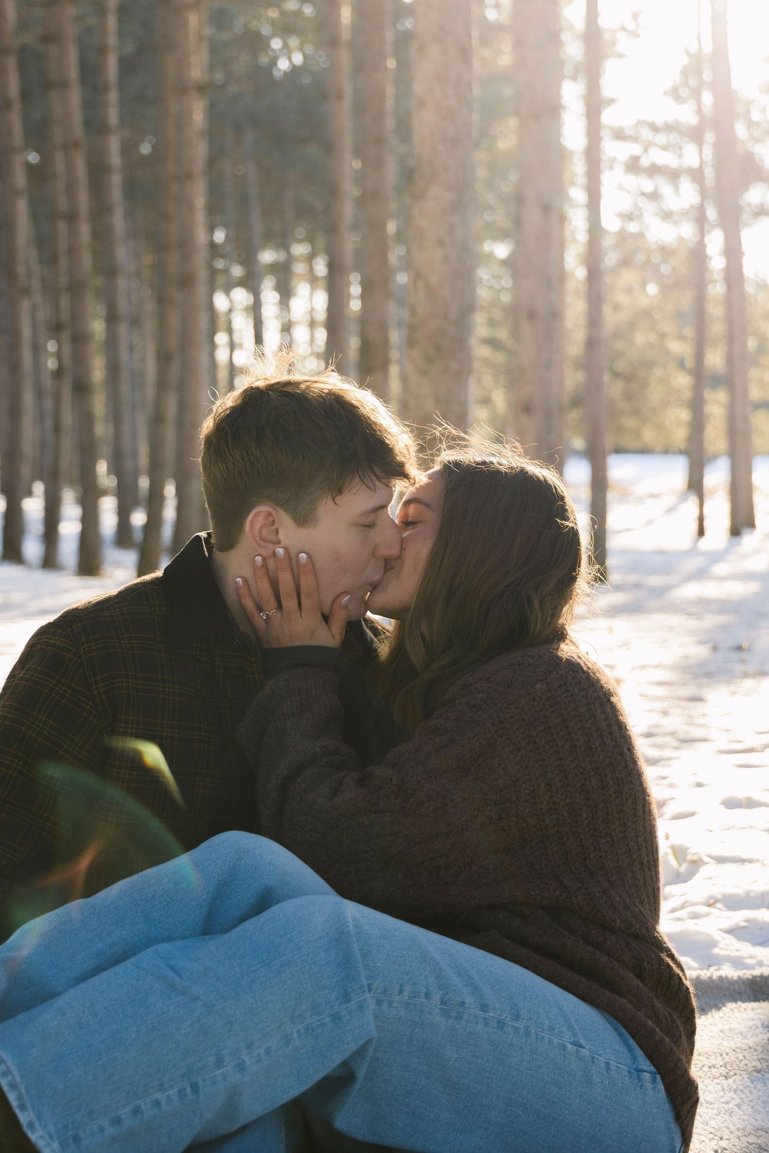 Romantic winter scene of couple embracing in snowy forest with sunlight streaming through trees.