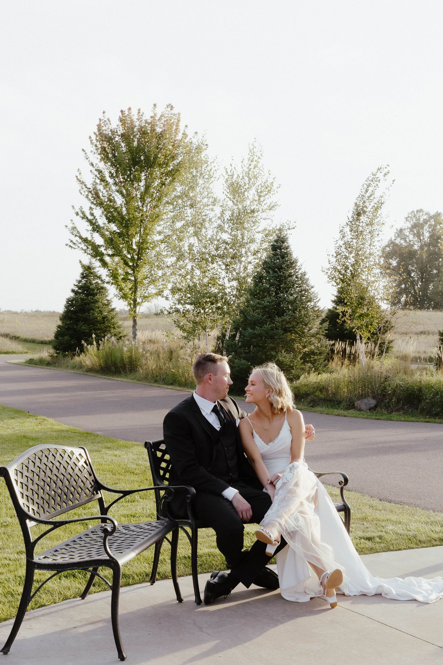 A couple in formal wedding attire sits together on outdoor benches at sunset.