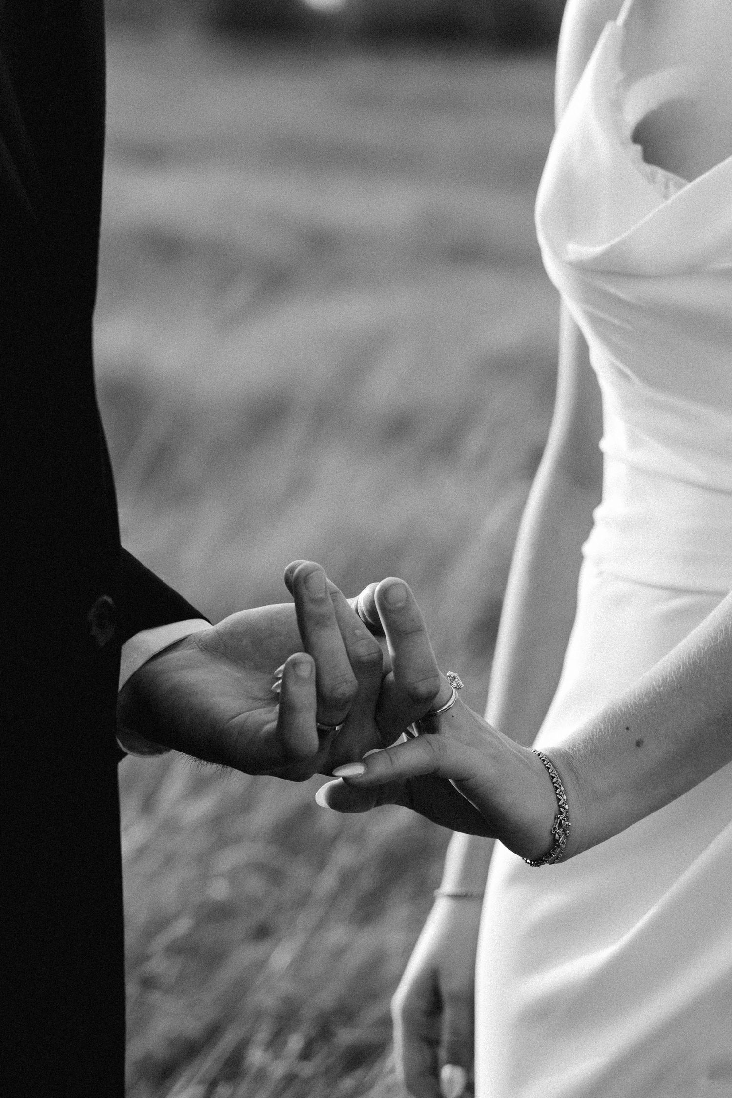 Black and white photo of bride and groom holding hands, showing wedding rings, bracelet, and elegant white dress.