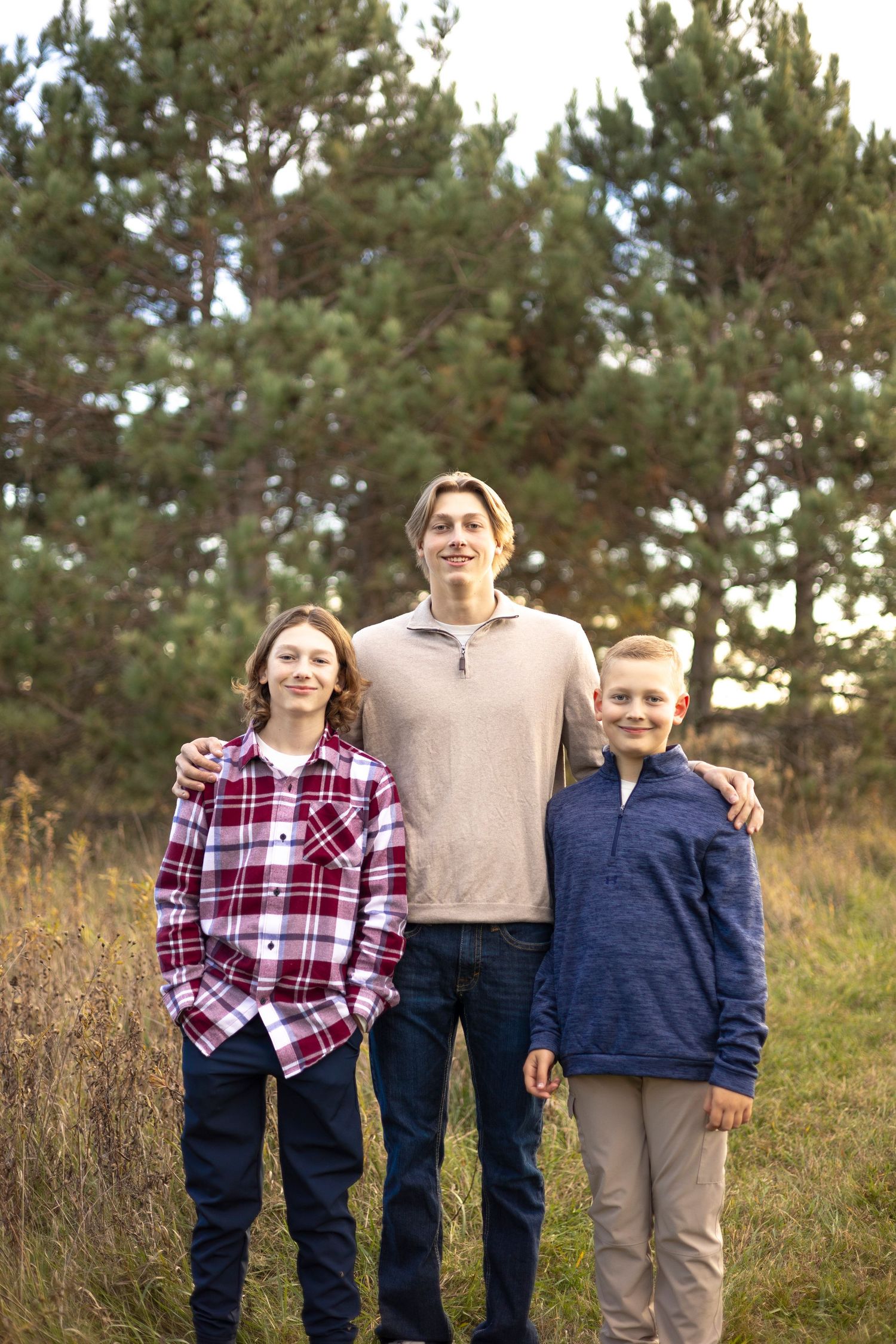 Three people pose together outdoors wearing coordinated casual outfits against pine trees.