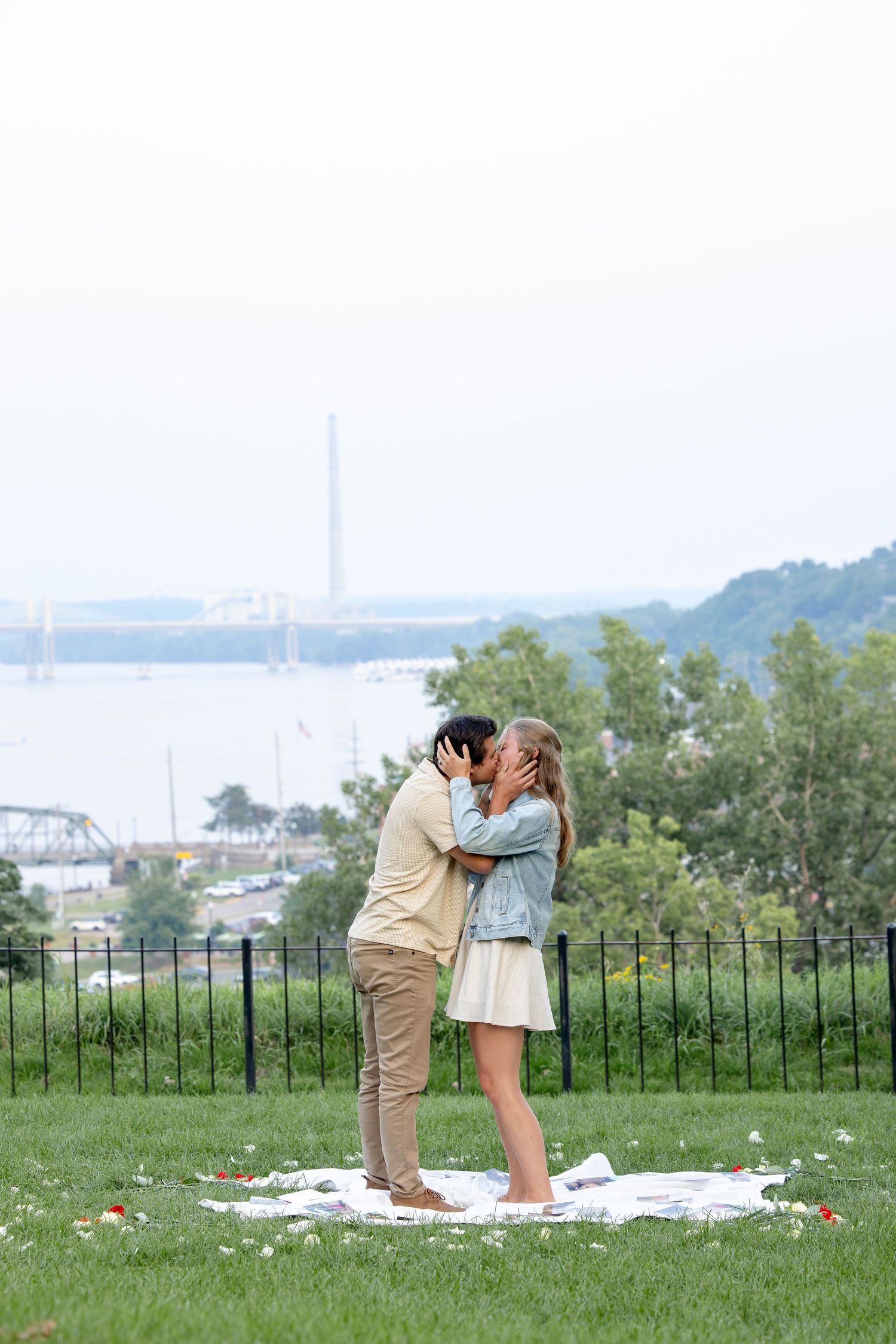 A couple shares a romantic moment on a picnic blanket with a bridge visible in the background.