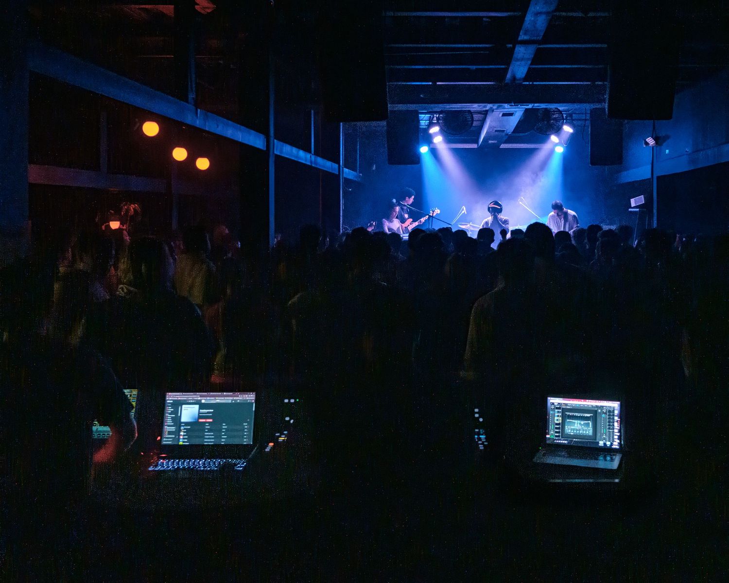 Dark concert venue with blue stage lighting and crowd silhouettes in foreground.