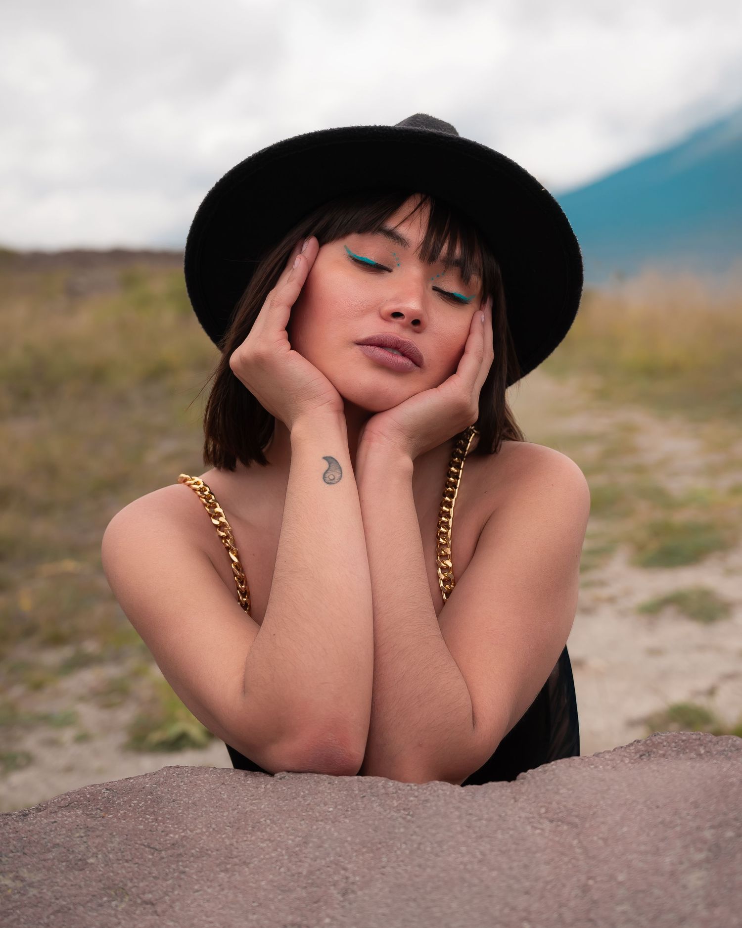Artistic portrait with black hat and dramatic shadows against a natural landscape.