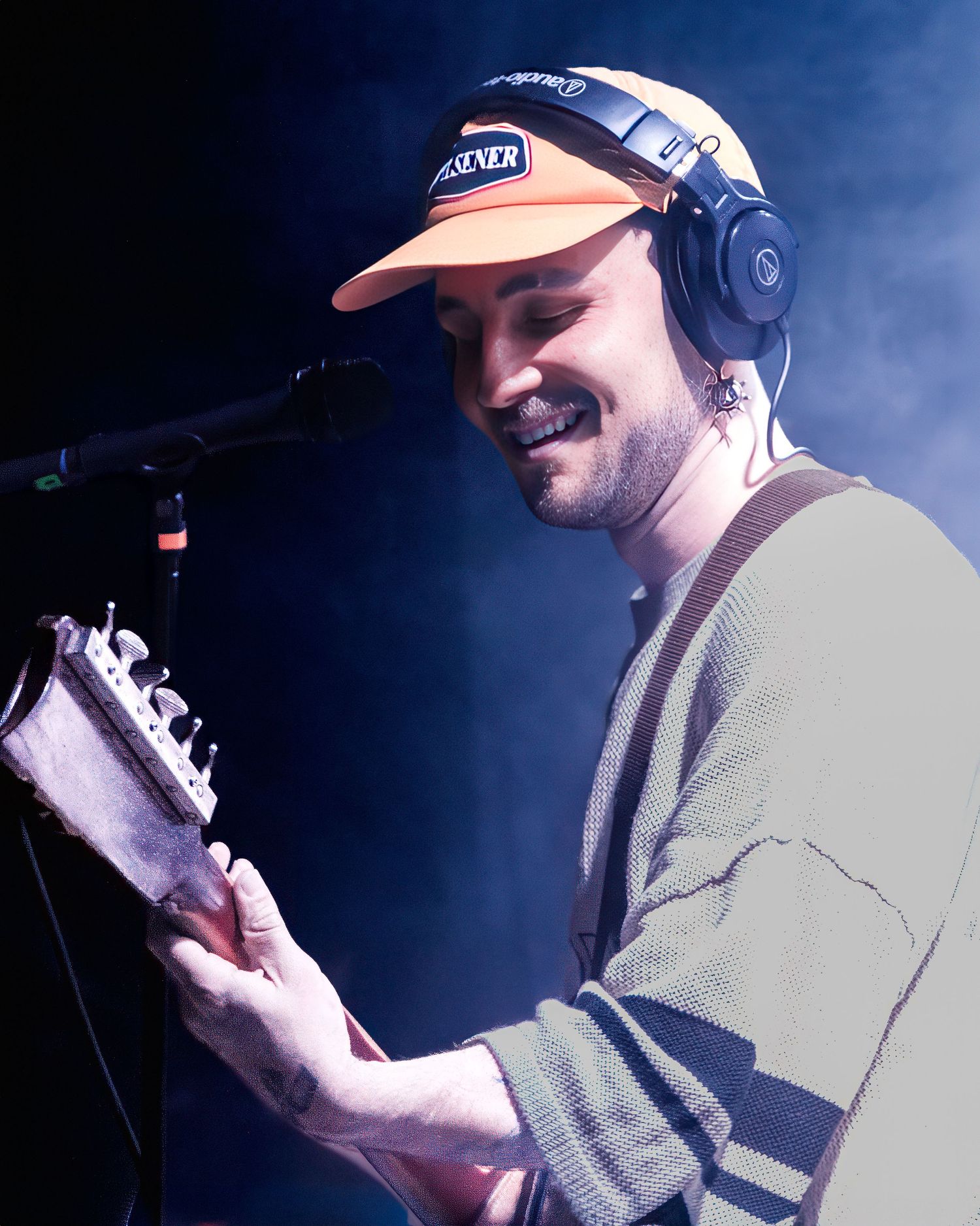 Concert performer with cap and headphones singing into microphone under blue stage lights.