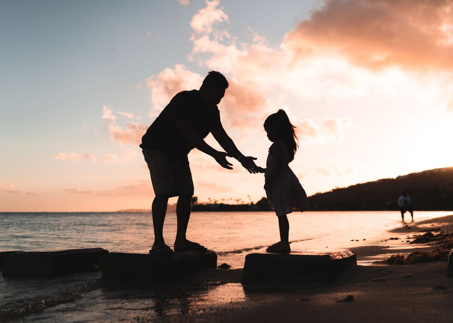 Two silhouettes share a tender moment on rocky coastal outcrop during sunset.
