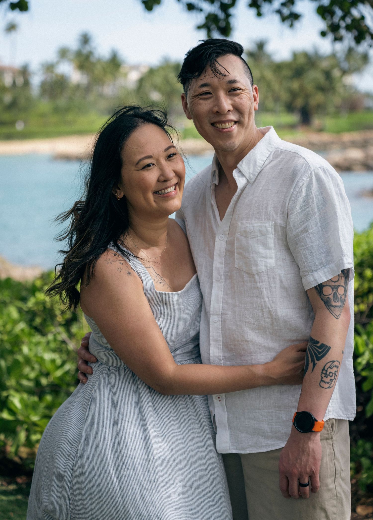A smiling couple embraces outdoors near a tropical beach, woman in gray dress, man in white linen shirt with tattoos.