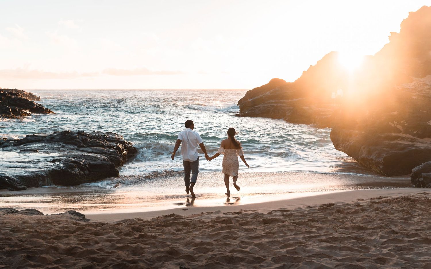 Silhouettes of people walking on beach at golden hour with waves and rocky cliffs in Hawaii.