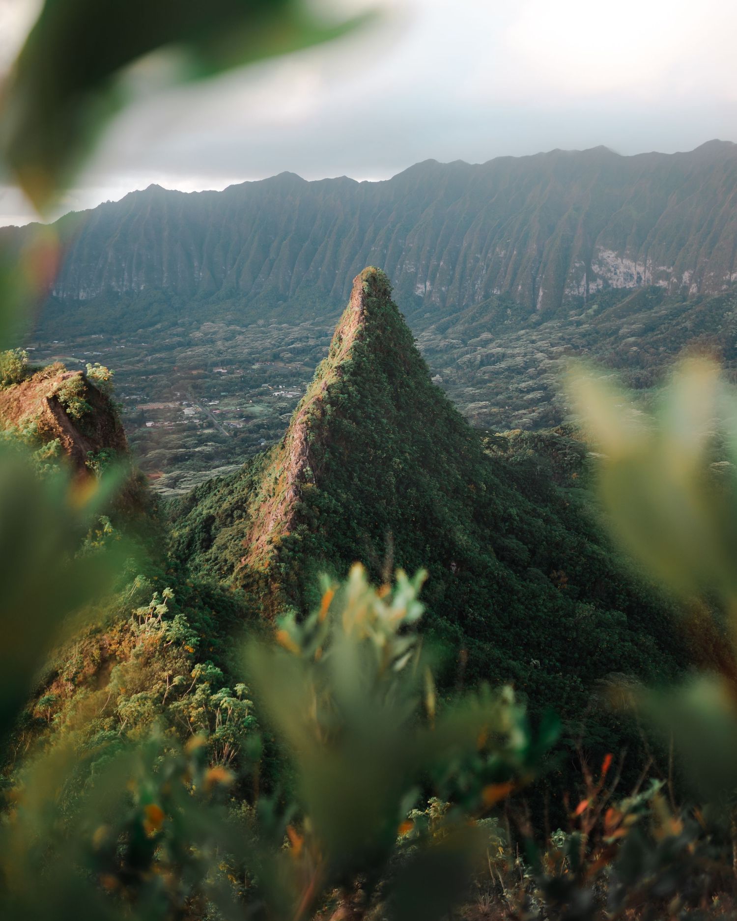 Dramatic mountain ridges and peaks rise against a misty sky in a lush tropical landscape.