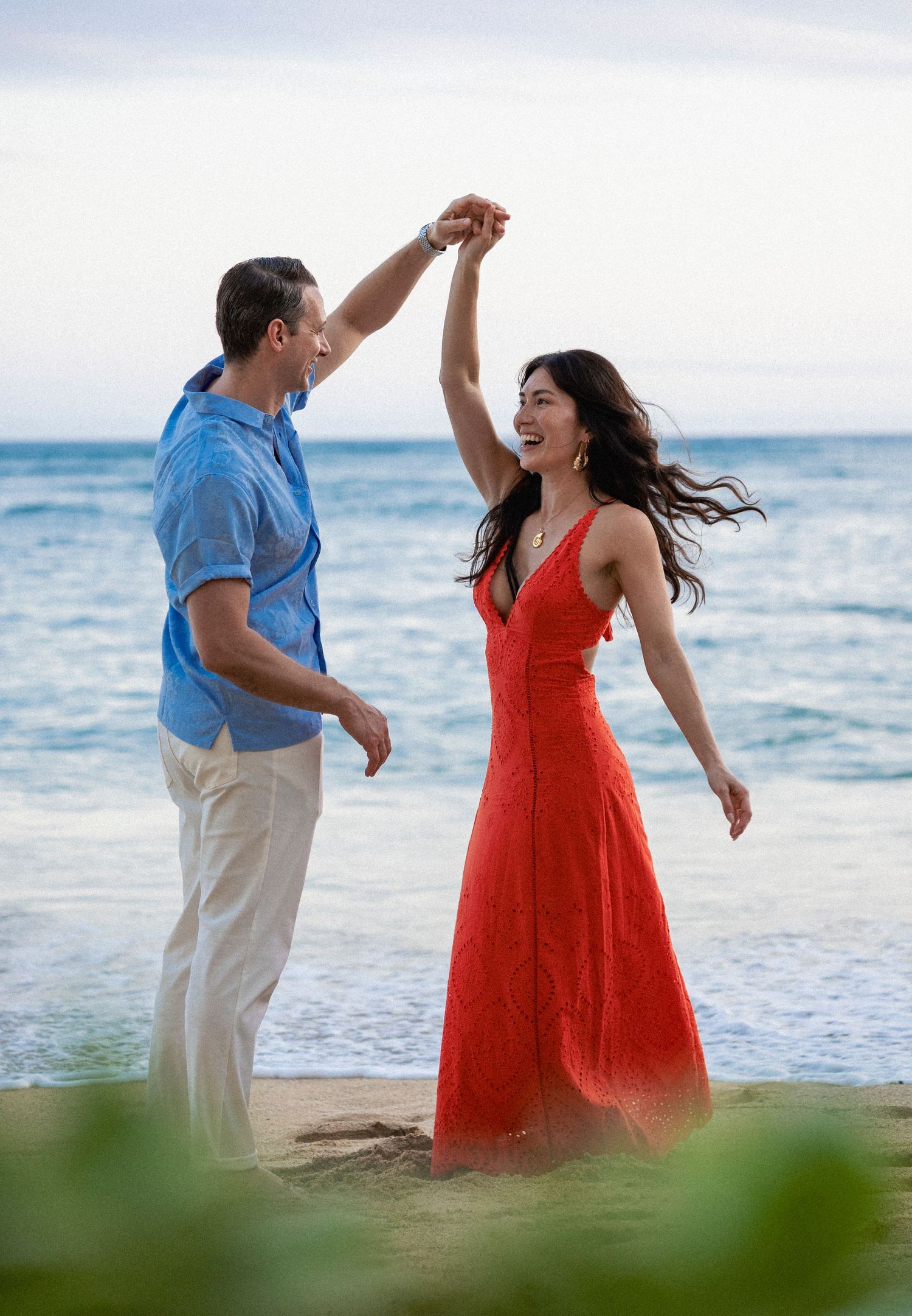 Couple dancing on a beach, woman in red dress being twirled by man in blue shirt, ocean backdrop.