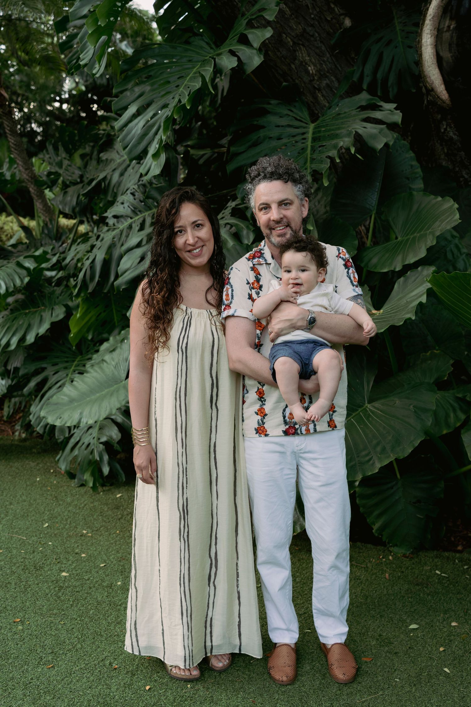 A family of three poses together against a backdrop of large tropical monstera leaves.