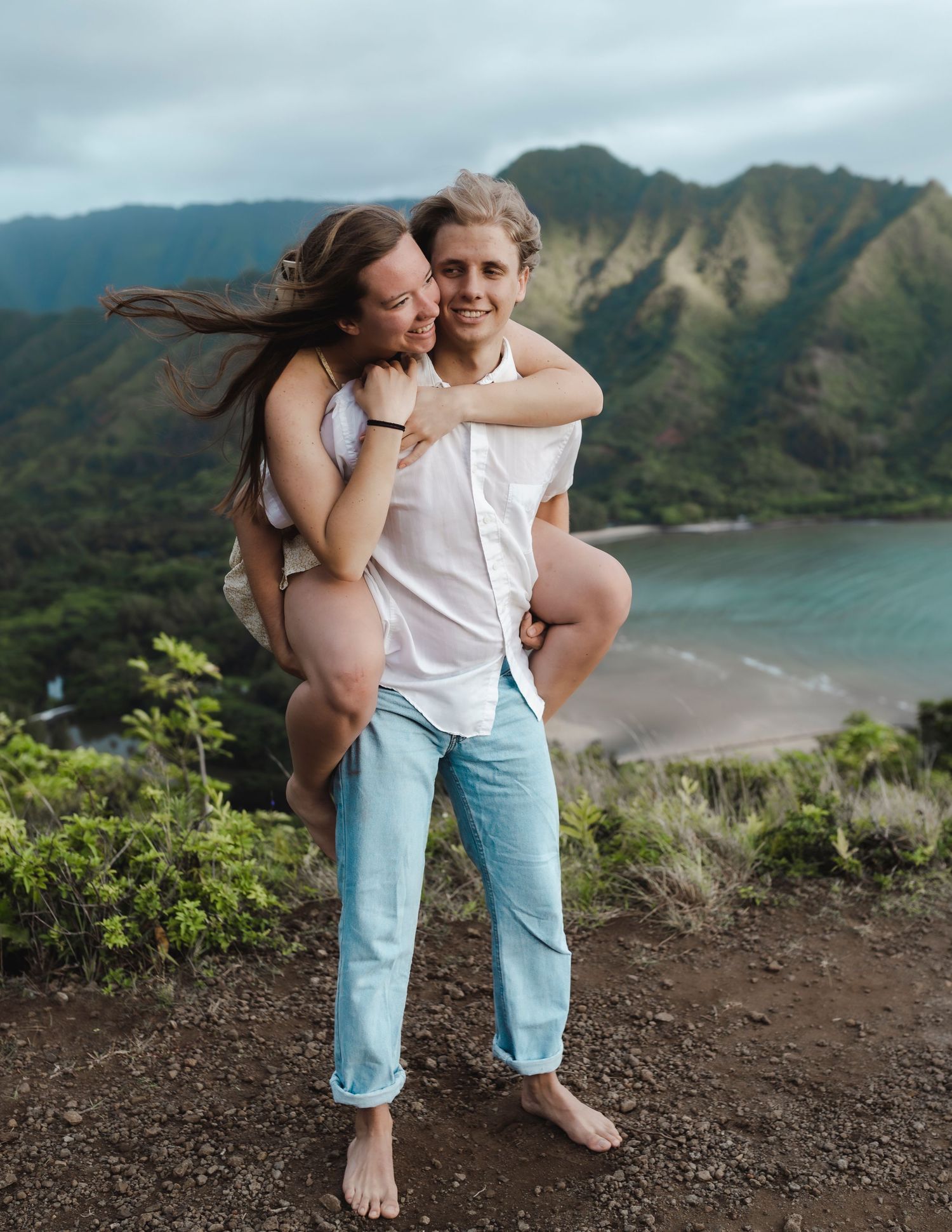 Couple posing on a scenic Hawaiian overlook, man giving woman a piggyback ride with lush mountains and beach behind.