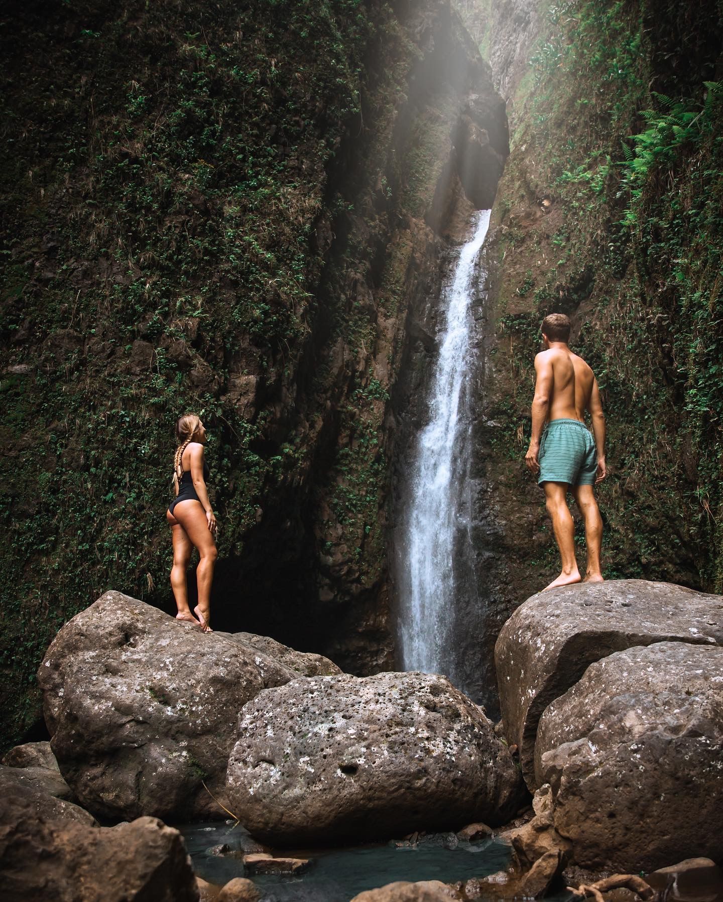 Couple standing on rocks admiring a lush tropical waterfall cascading down a mossy canyon wall.