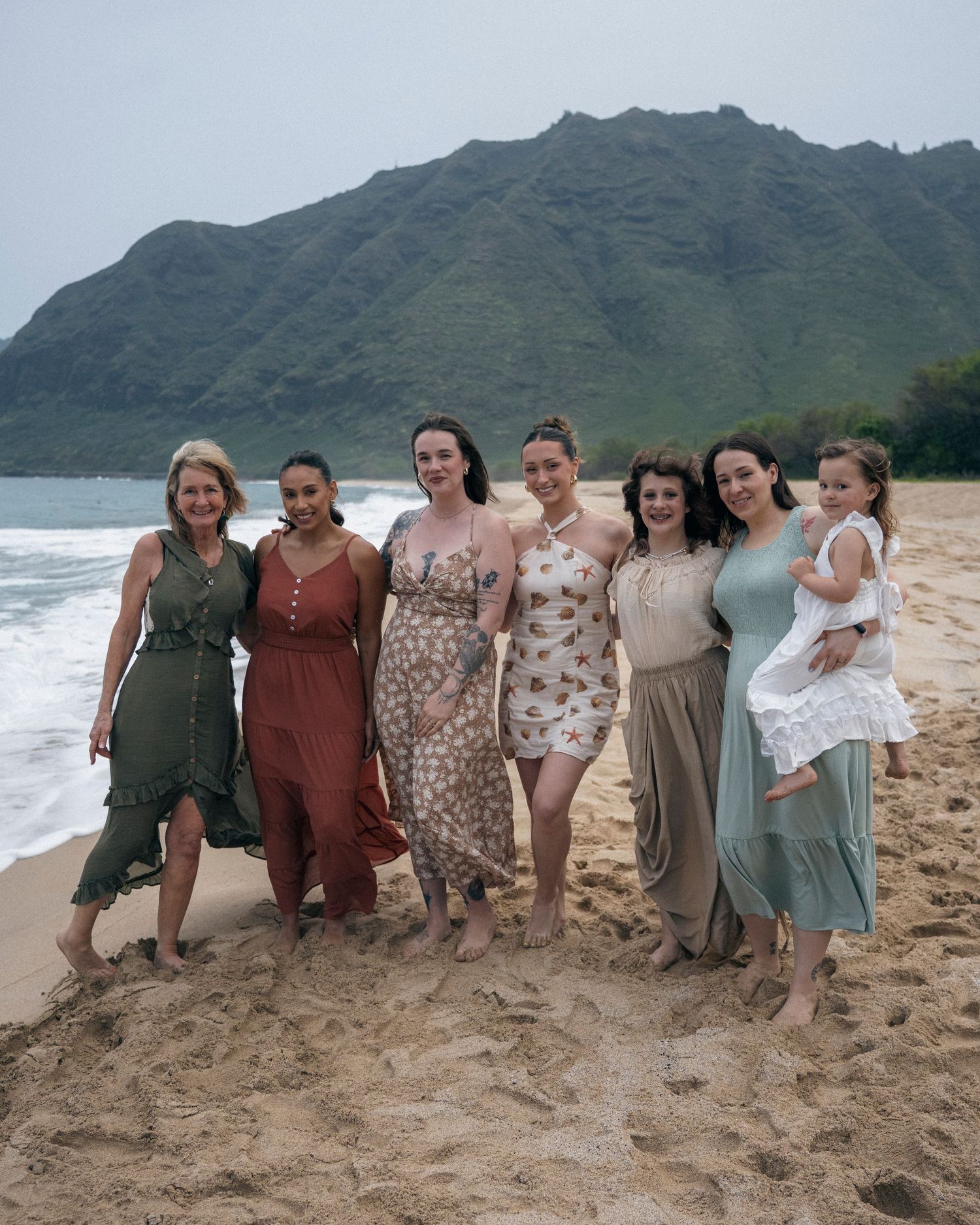 A group in coordinated earth-tone outfits poses together on a scenic beach with mountains in the background.