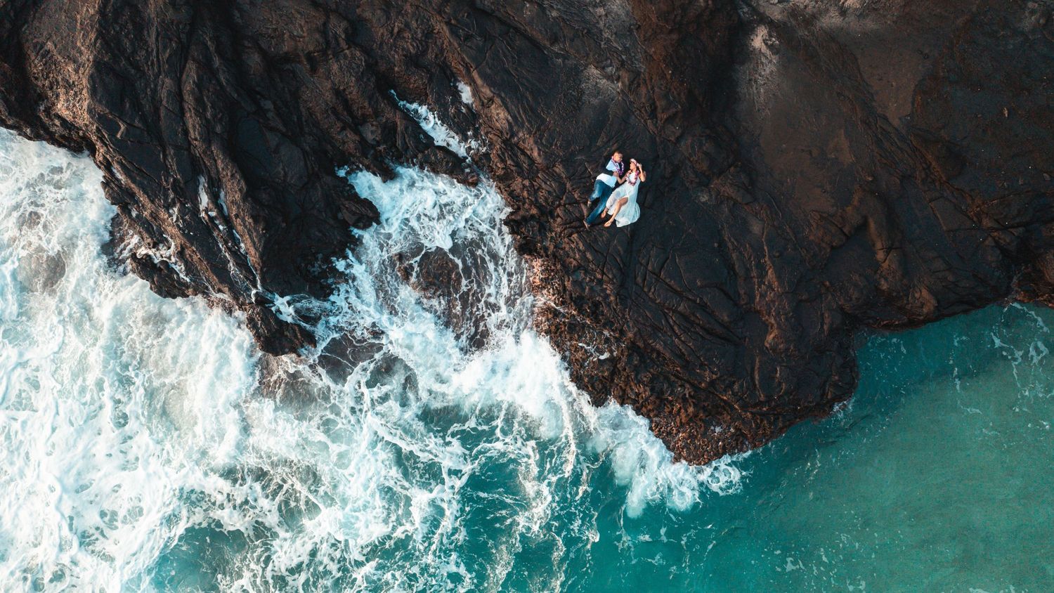 Dramatic aerial view of turquoise ocean waves crashing against dark volcanic rocks.