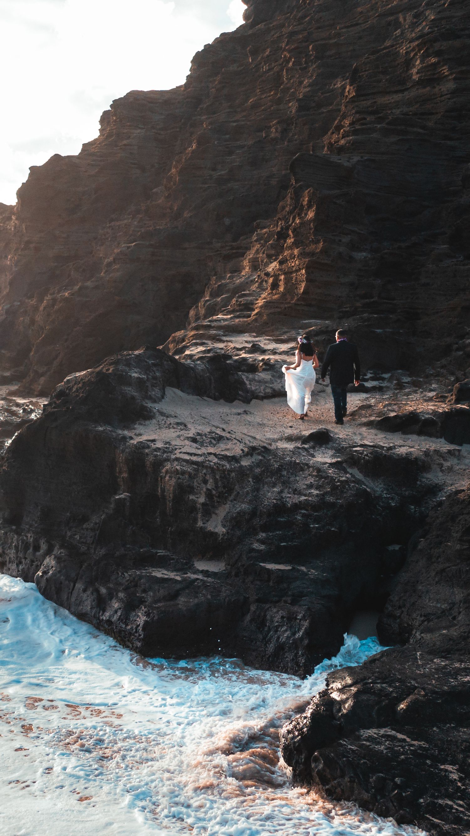 Couple in wedding attire walking on rocky coastal cliffs with turquoise waves crashing below during golden hour.