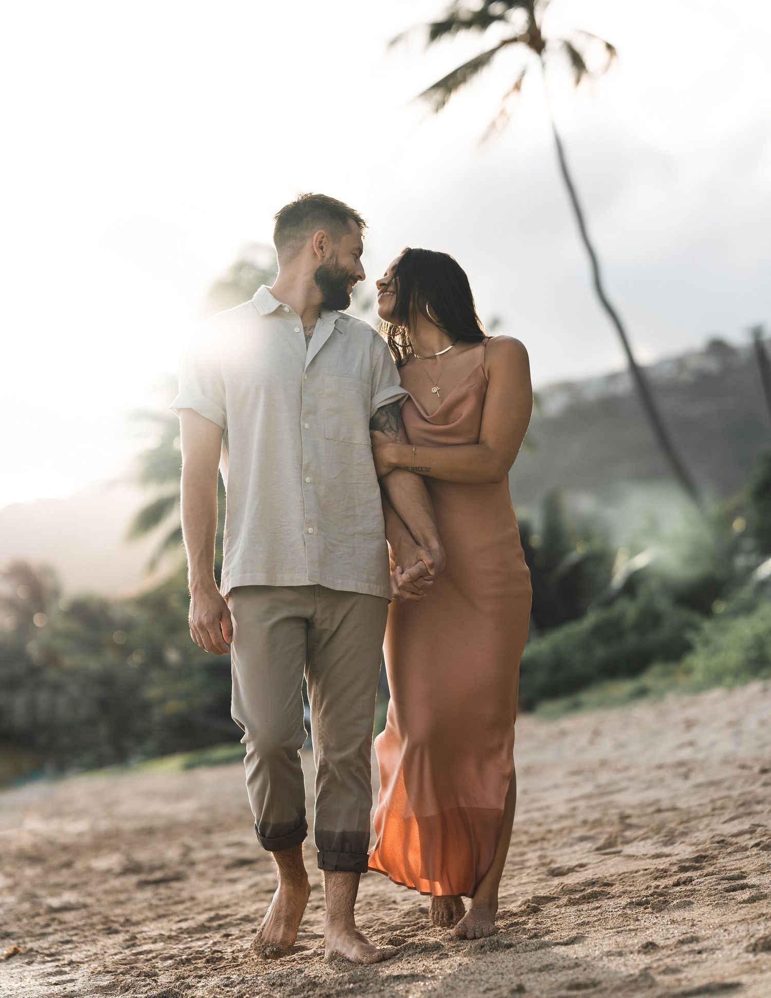 Romantic couple strolls along a sandy beach at sunset with palm trees in the background.