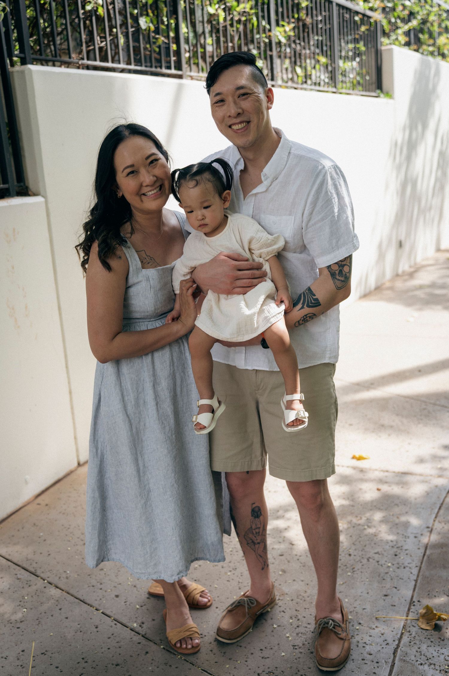 A family poses together in casual summer attire against a white wall with greenery.