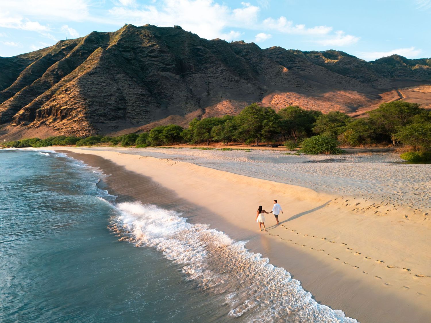 Aerial view of a couple walking on a secluded sandy beach with turquoise waves and dramatic green mountains backdrop.