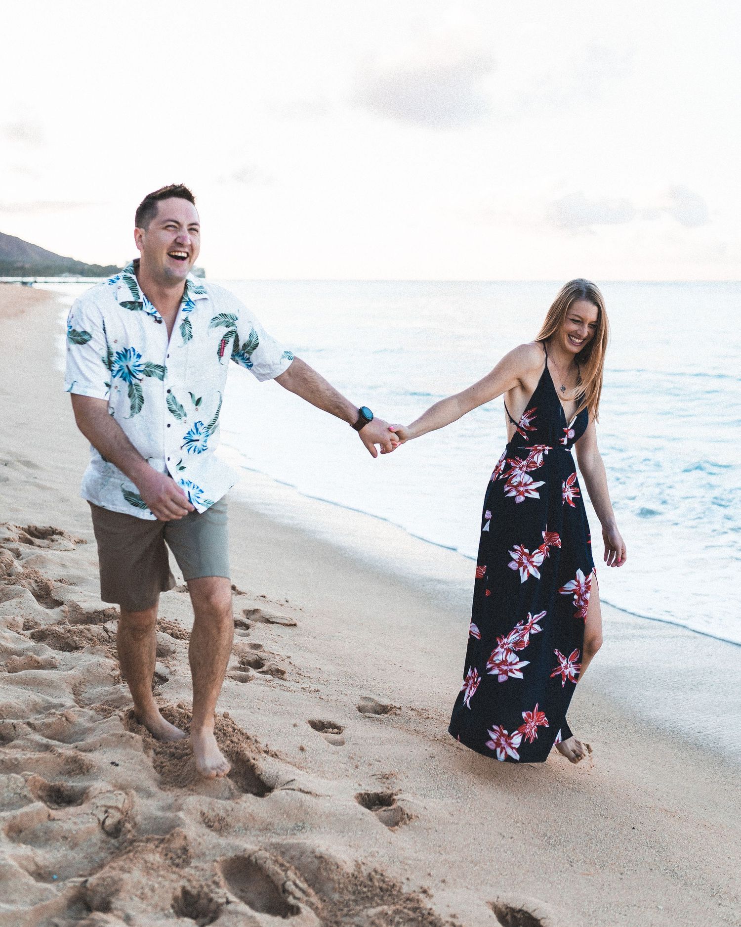 Couple holding hands and laughing while walking barefoot on a sandy beach, wearing floral outfits near the ocean.