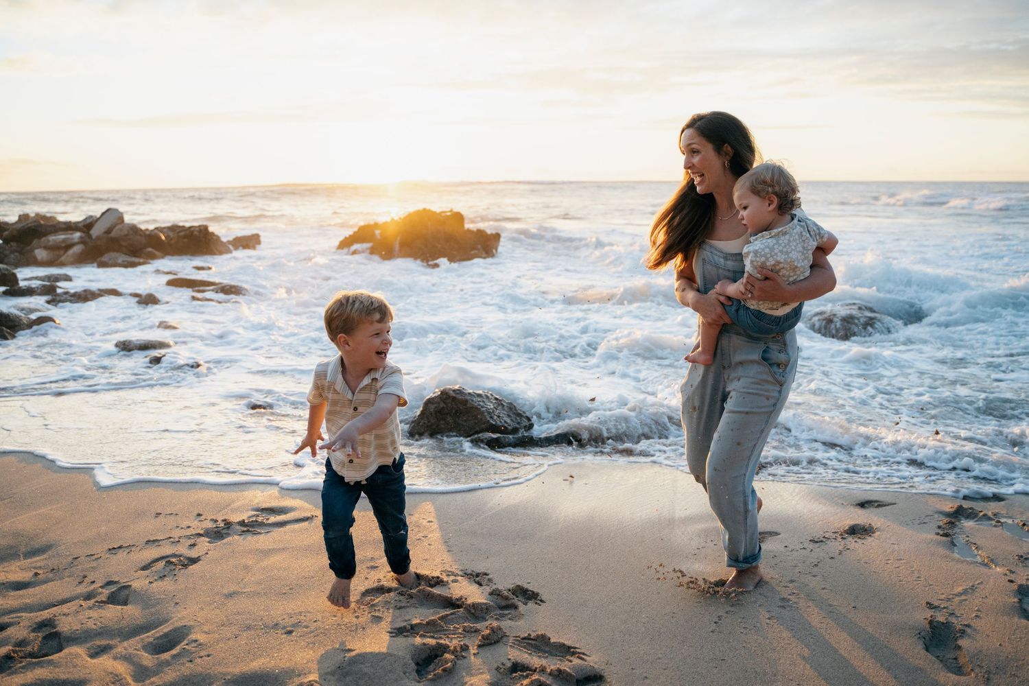 A mother holds her baby while her toddler plays at the beach during a golden sunset with waves crashing on rocks.