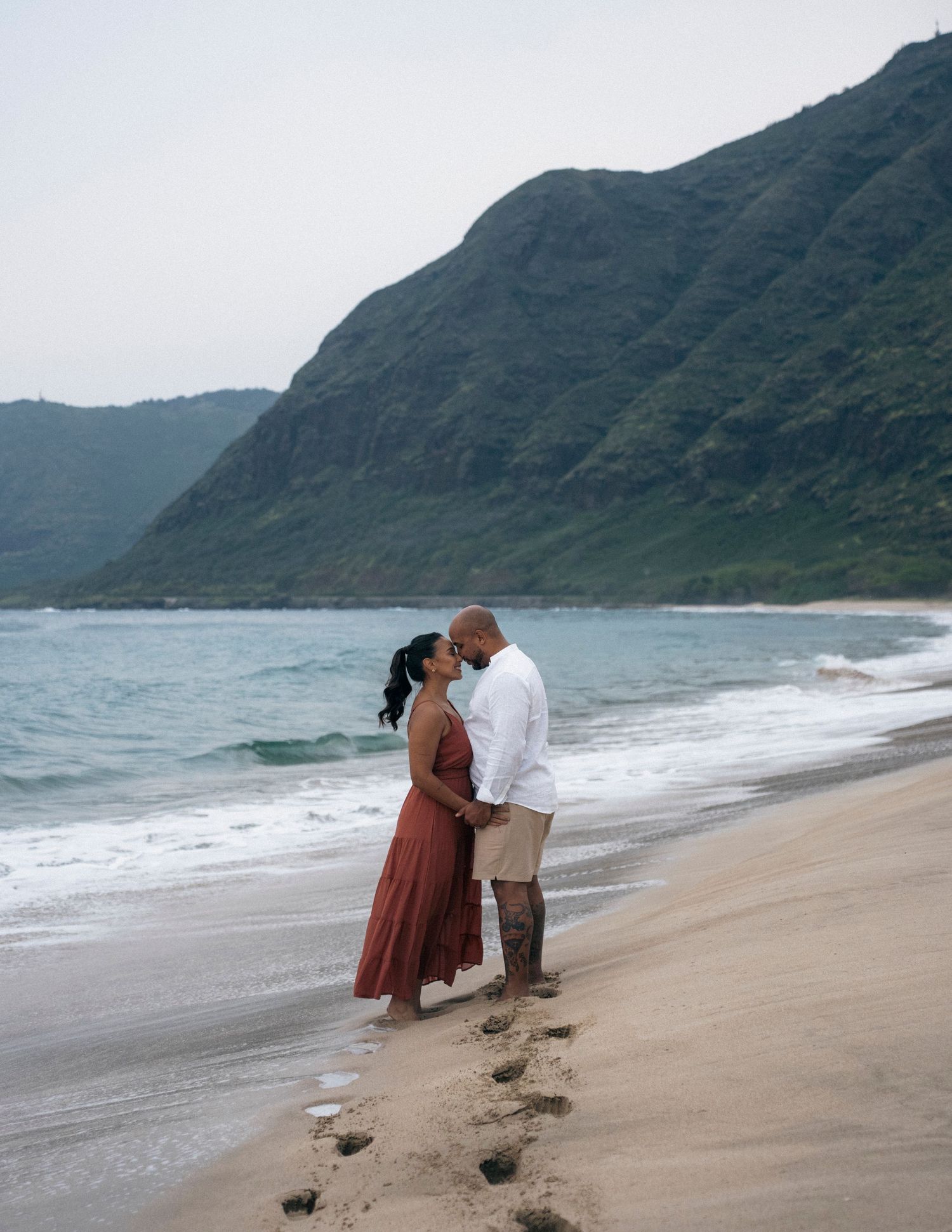 Couple sharing a romantic moment on a sandy beach with lush green mountains and ocean waves in the background.