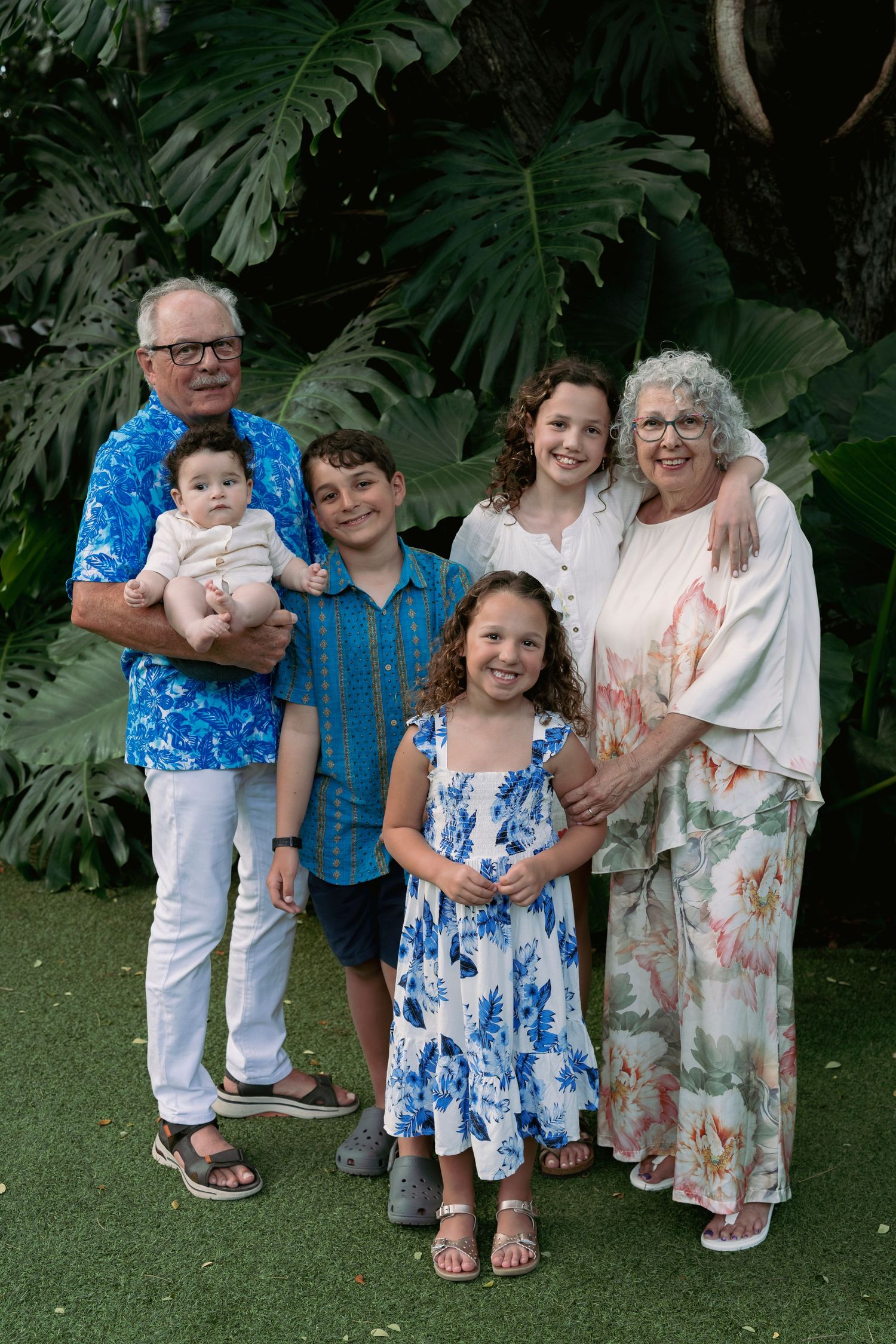 A multi-generational family poses together against a backdrop of large tropical monstera leaves.