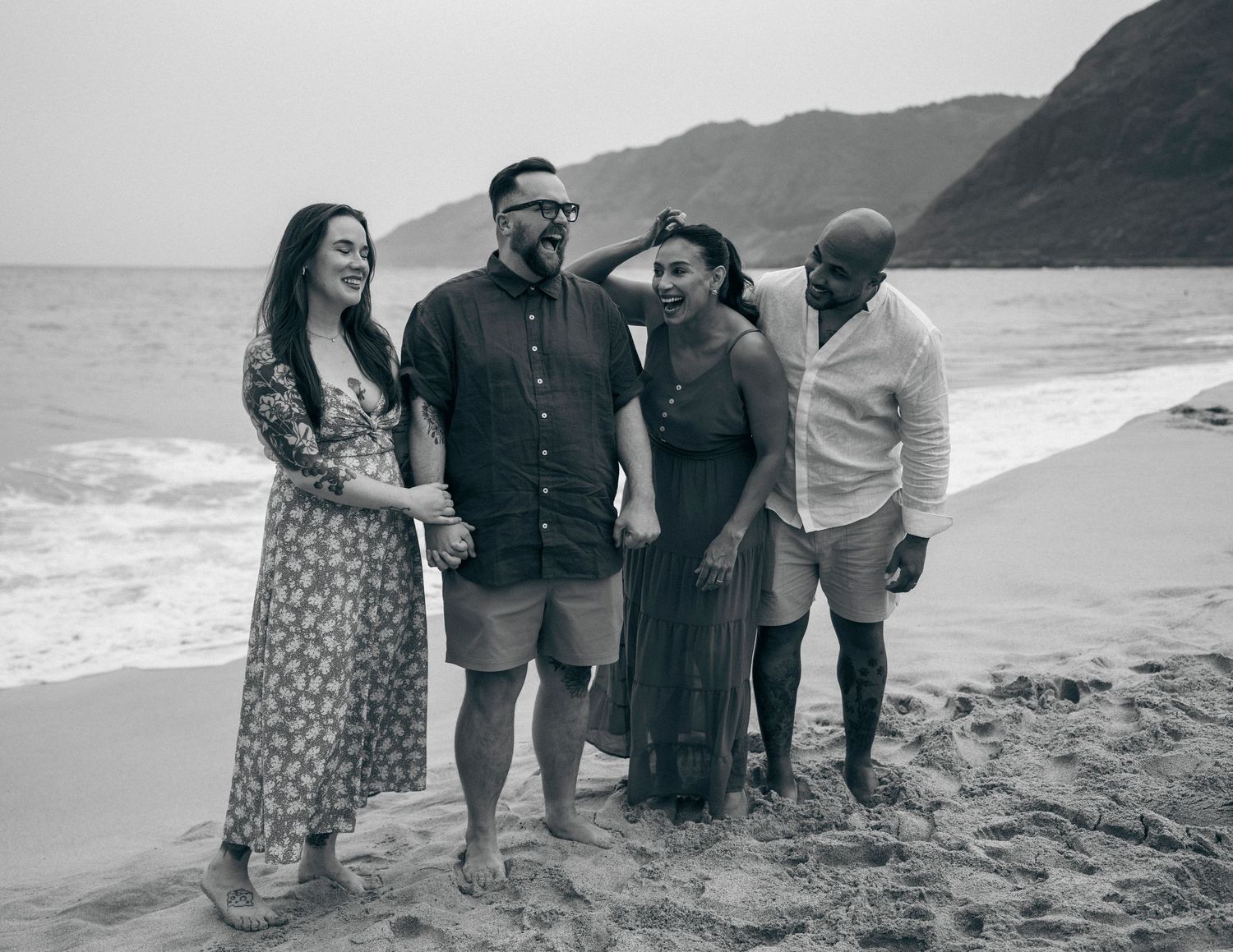 Four friends laughing together on a beach with ocean waves and mountain backdrop in black and white photo.