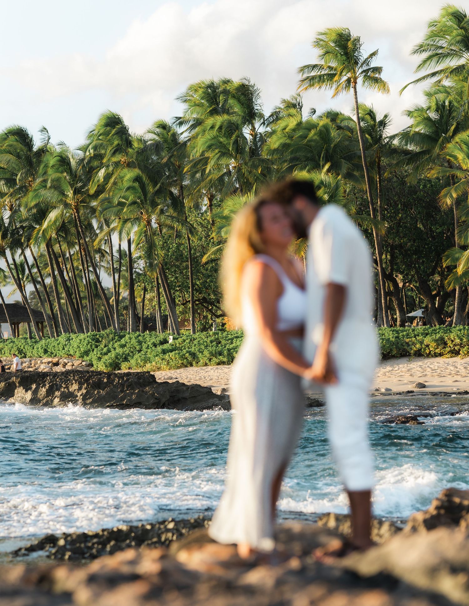Couple embracing on a Hawaiian beach with palm trees and crashing waves at sunset.