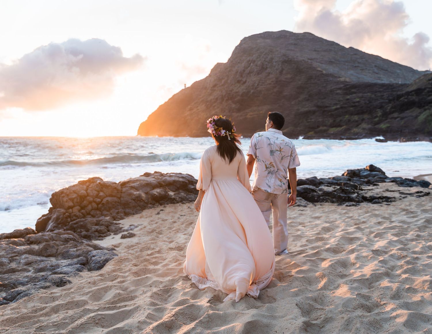 A couple in wedding attire walks hand-in-hand on a sandy beach at sunset with rocky cliffs and ocean waves.