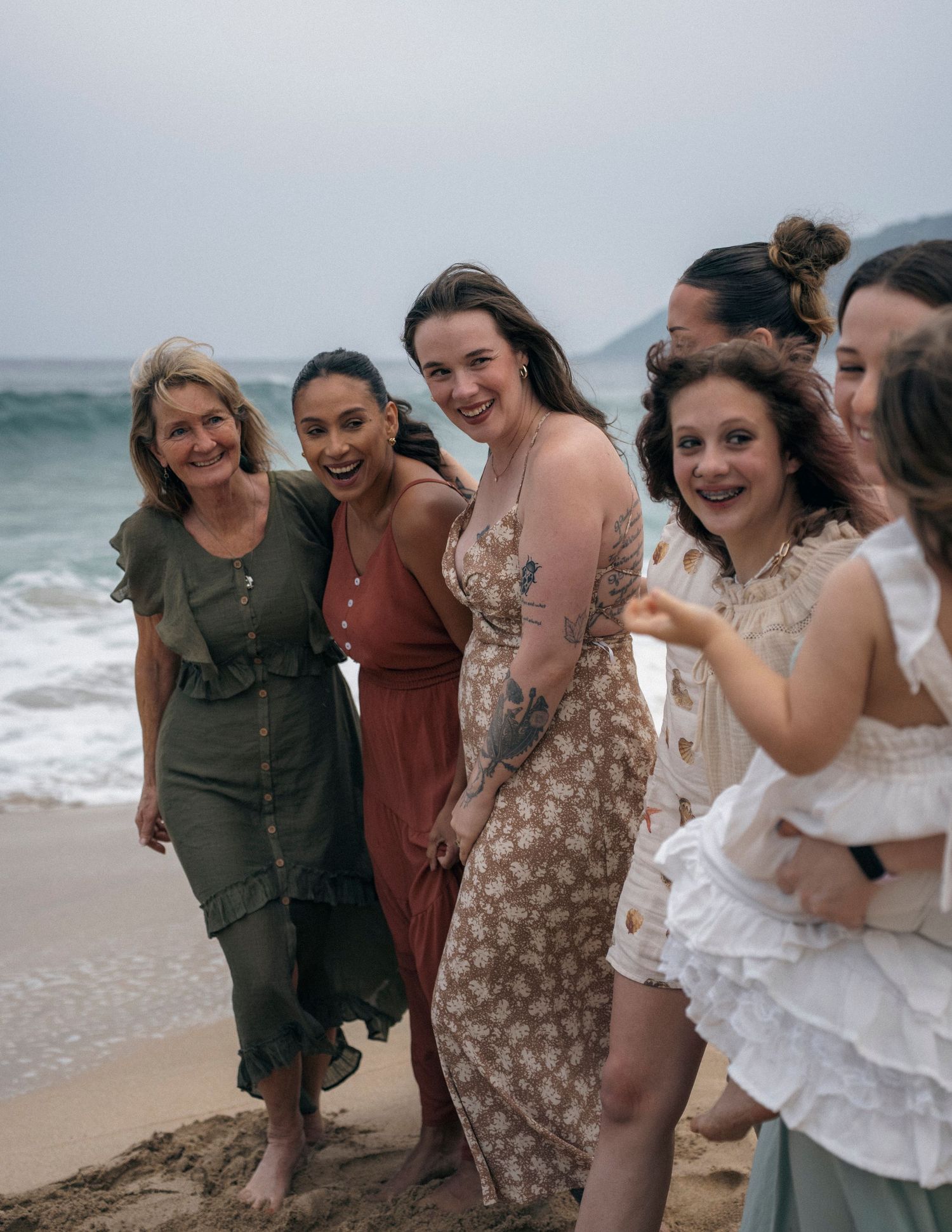 Friends laughing and having fun together on a sandy beach with waves and mountains visible.