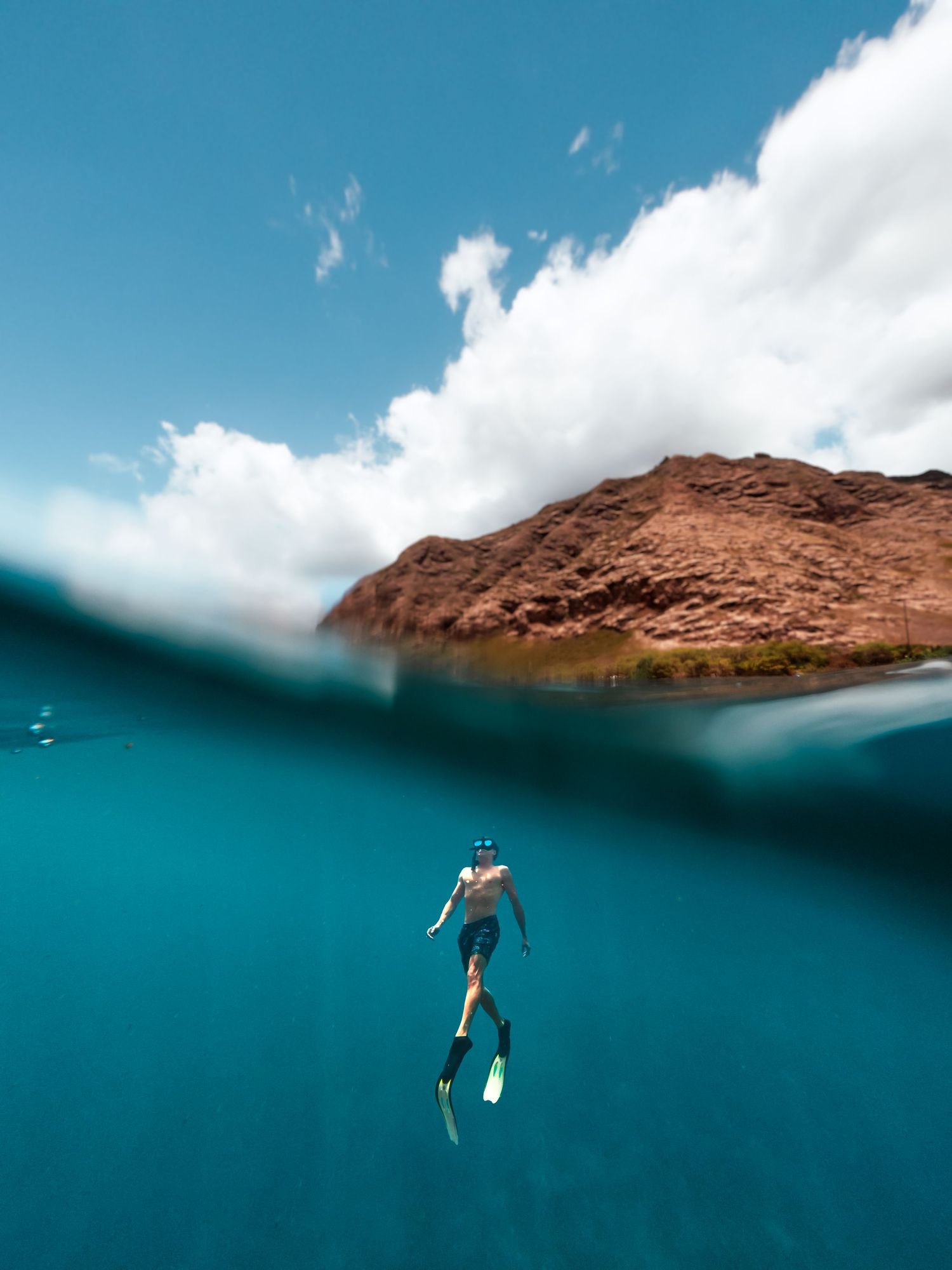 Split-shot underwater photography showing a snorkeler in turquoise waters against red rock formations.