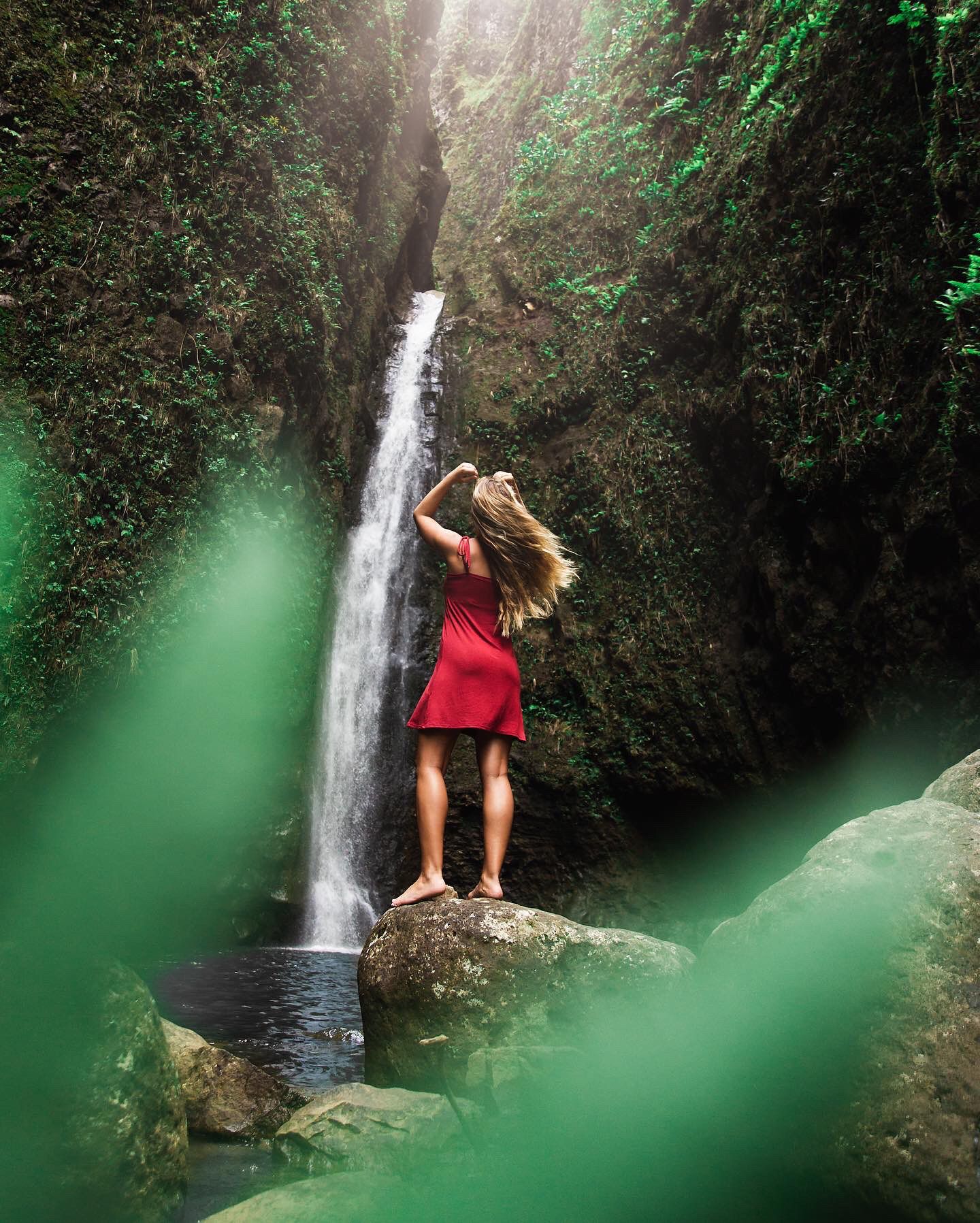 Woman in red dress standing on rock formation near cascading waterfall in tropical setting.