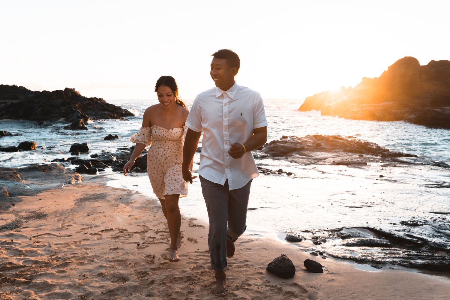 A couple walks hand in hand on a rocky beach at sunset with ocean waves and cliffs in the background.
