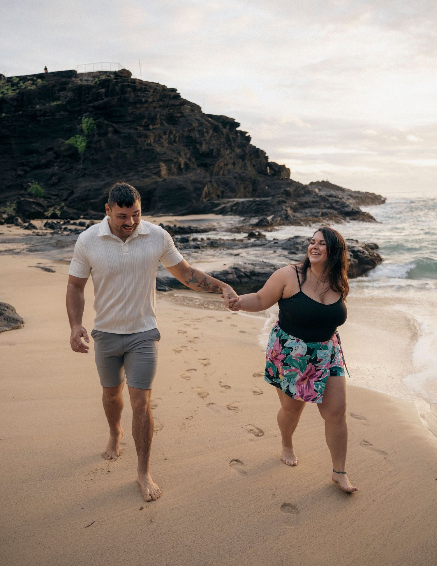 Couple holding hands and walking barefoot on a sandy beach with rocky cliffs and ocean waves at sunset.
