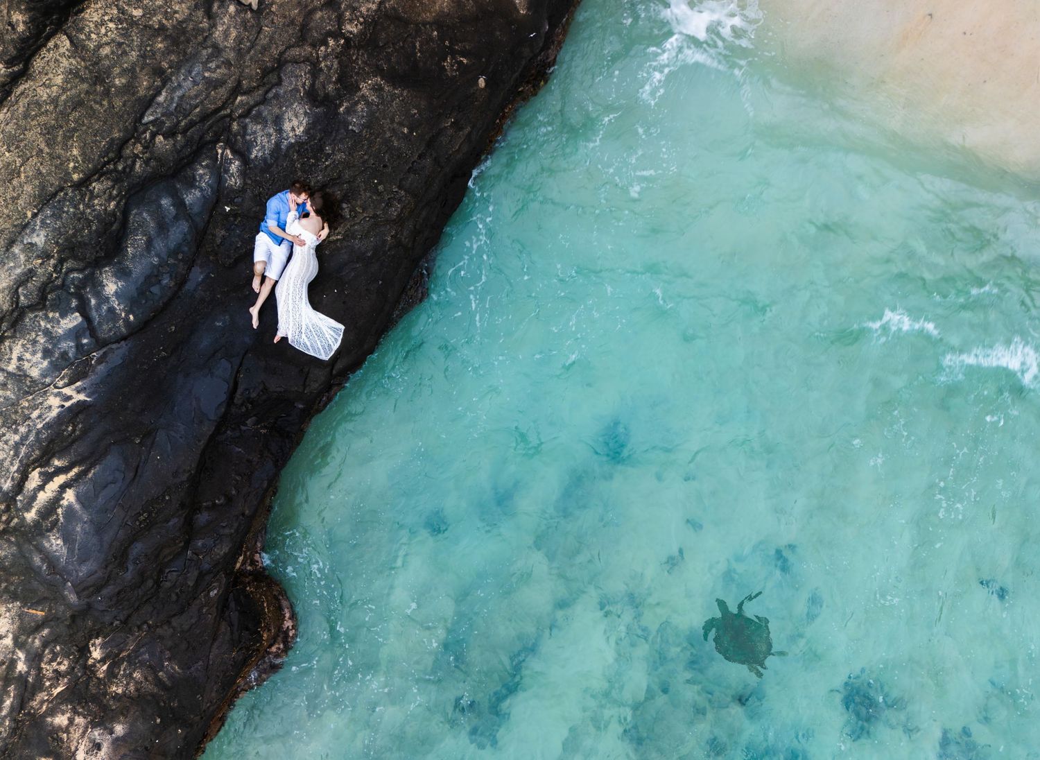 Aerial view of a wedding couple embracing on rocky coastal cliffs beside crystal clear turquoise ocean waters.