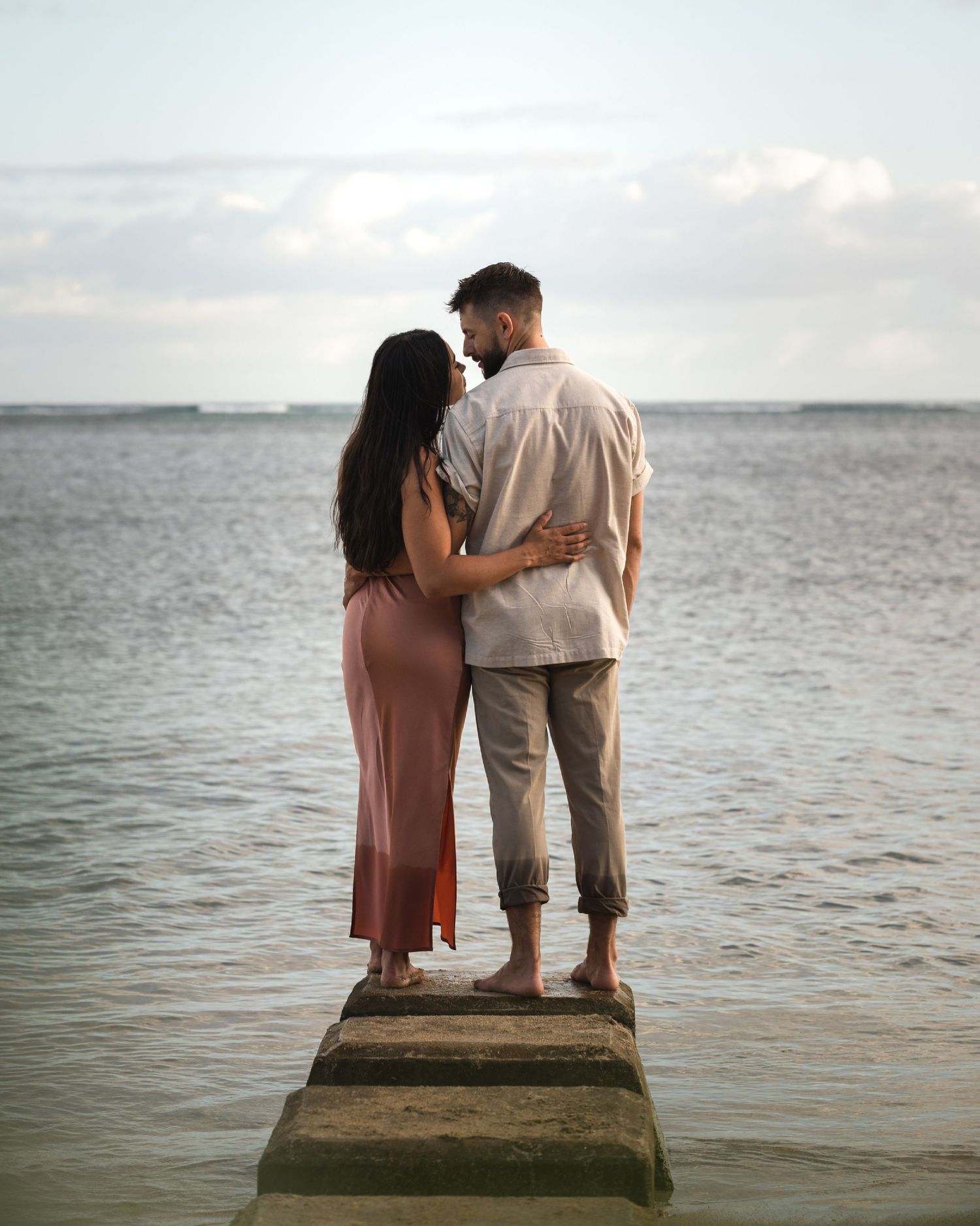 Couple embracing on a seaside pier at dusk, standing barefoot over calm ocean waters.