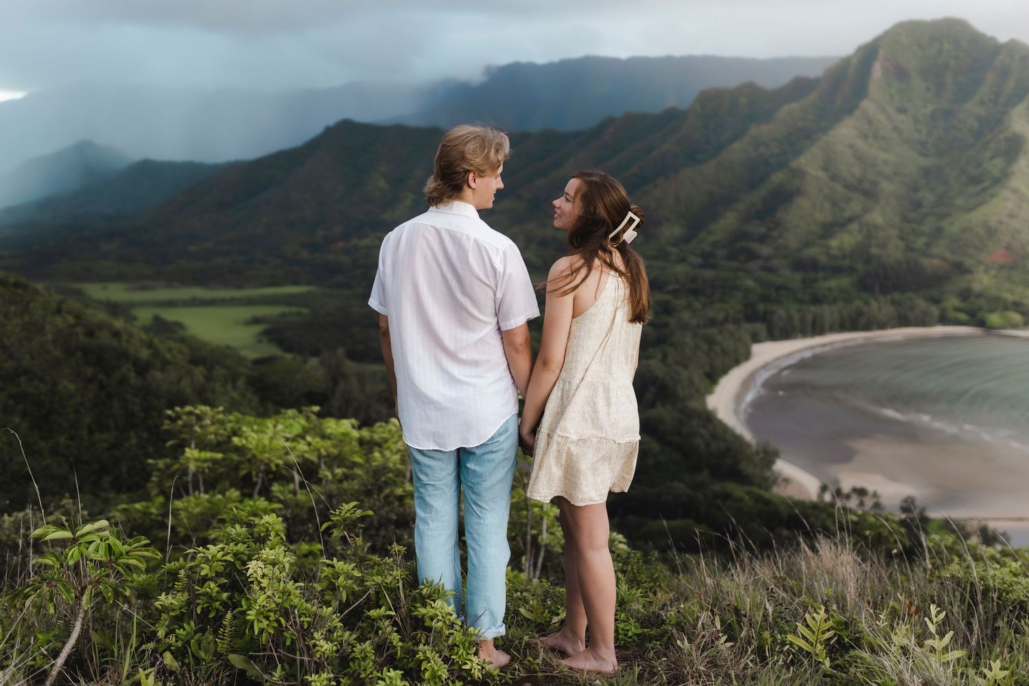 Couple holding hands overlooking lush green mountains and a scenic bay in Hawaii.