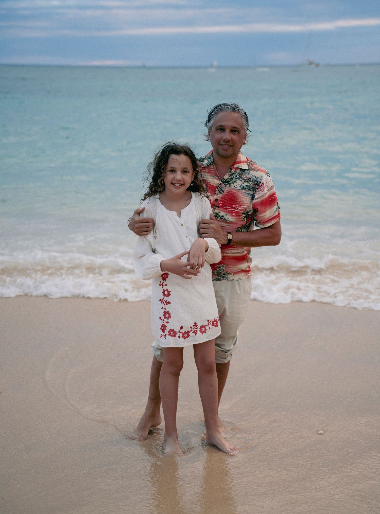 Two people stand together on a tropical beach wearing Hawaiian-themed clothing at the water's edge.