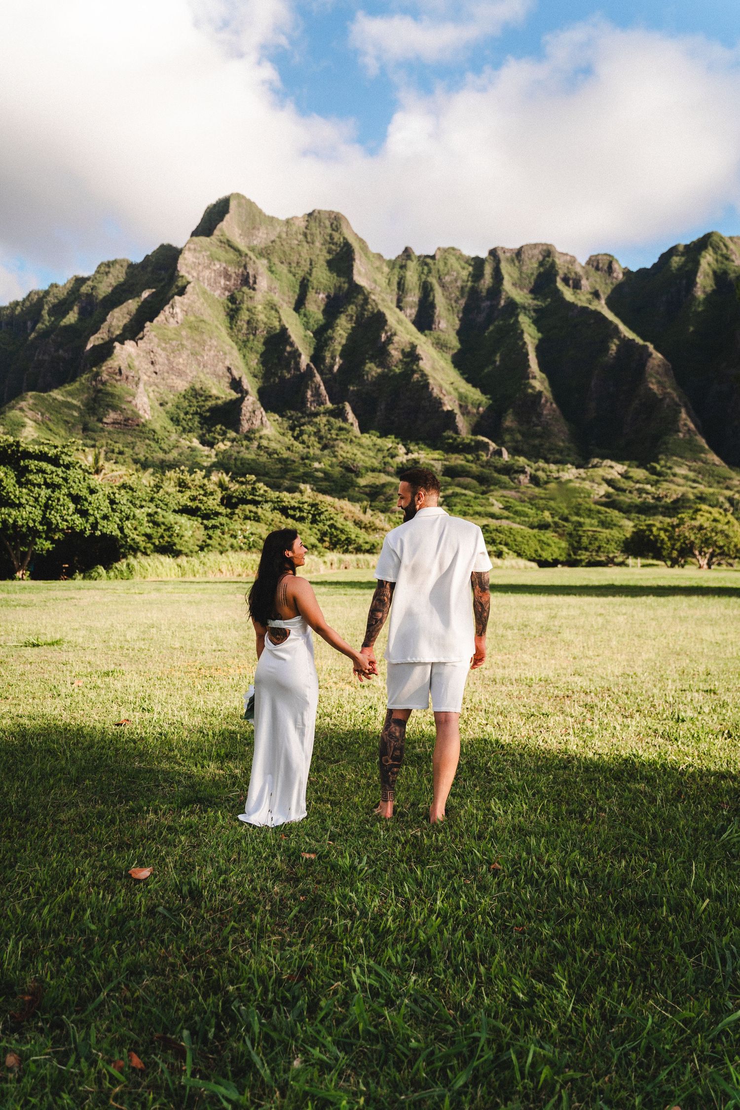 Couple in white outfits holding hands on green lawn with dramatic Ko'olau mountain range backdrop in Hawaii.