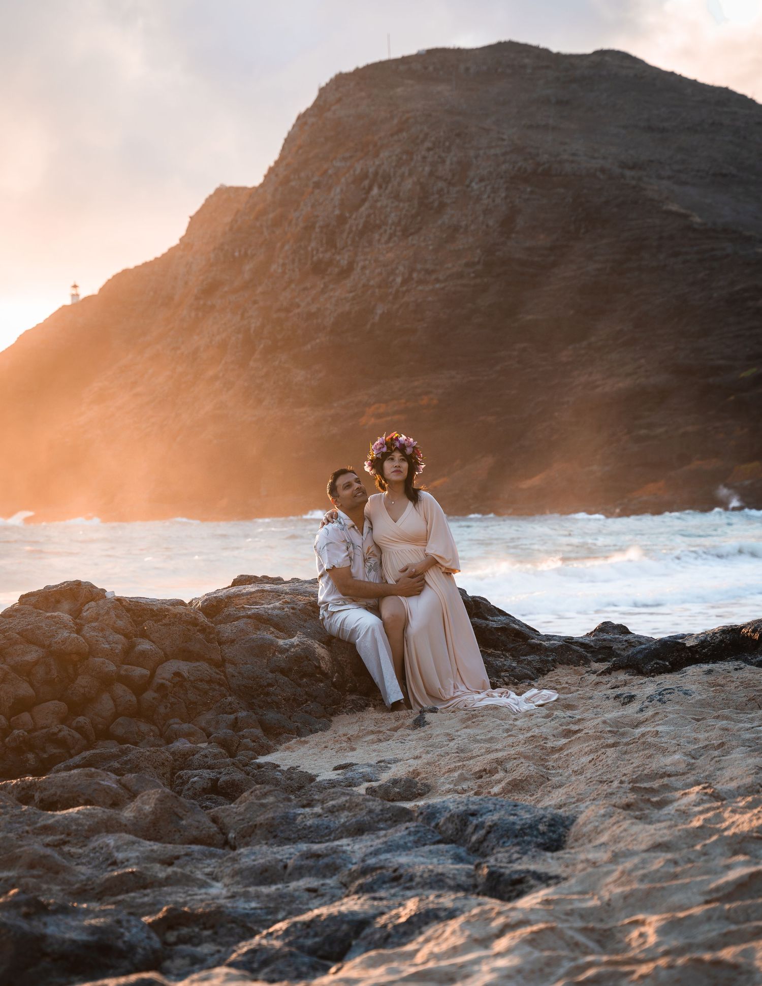 Couple sitting on rocky beach at golden sunset, woman in floral crown and flowing dress, dramatic cliffside ocean backdrop.